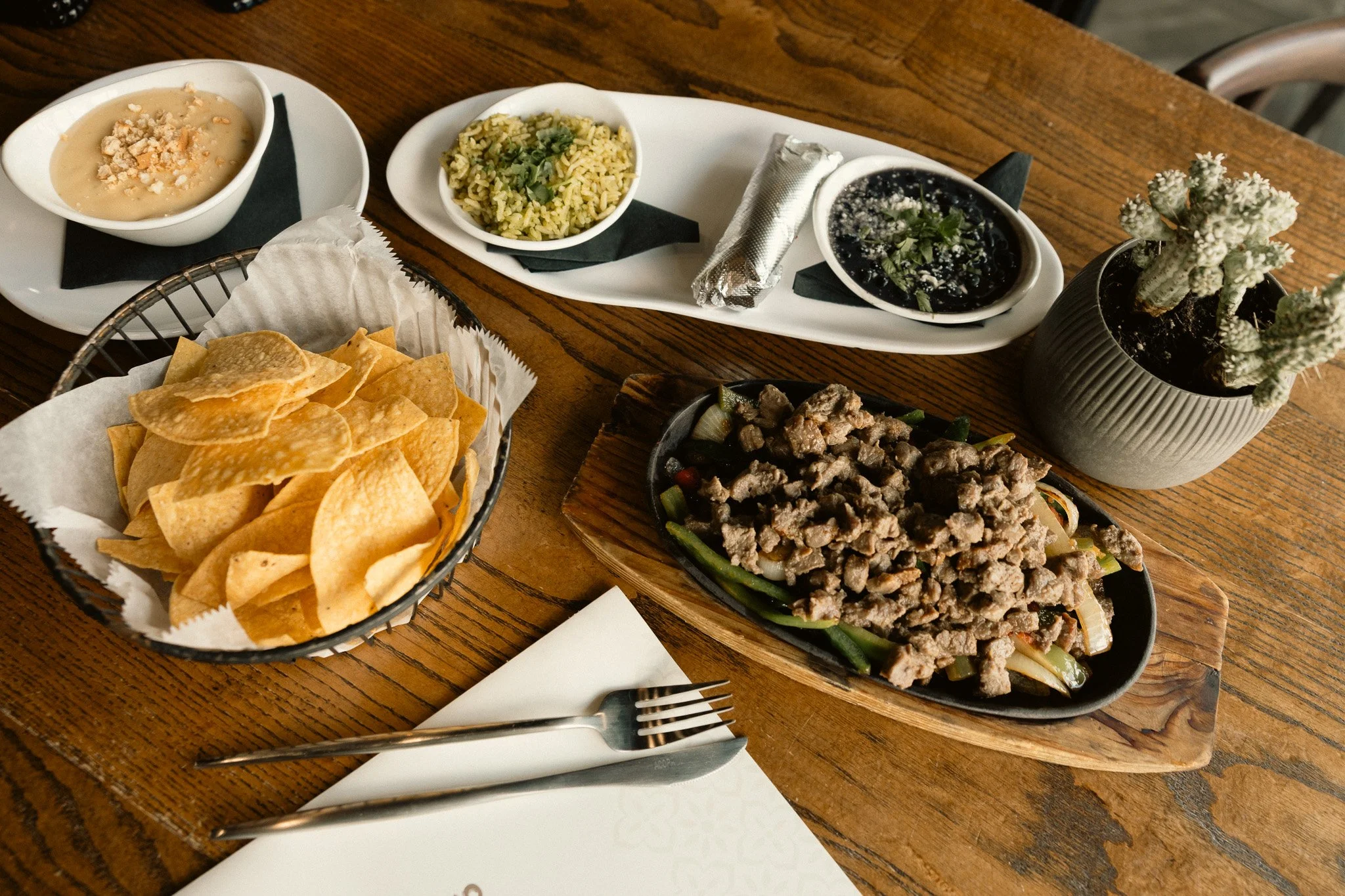 A wooden table with a basket of tortilla chips, a white oval plate with three small bowls of dips, falafel, ground beef stir-fry, and a potted cactus.