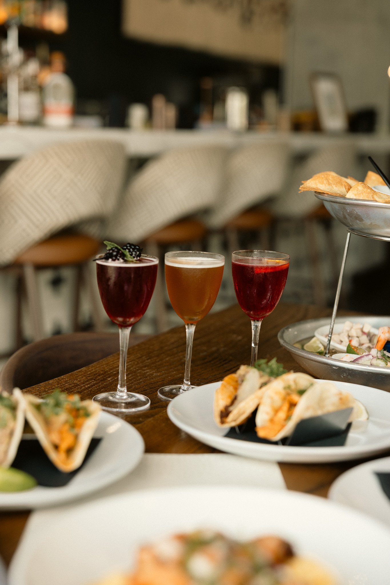 Three glasses of colorful beverages, possibly cocktails, on a wooden table, with plates of tacos and other snacks in a restaurant setting.