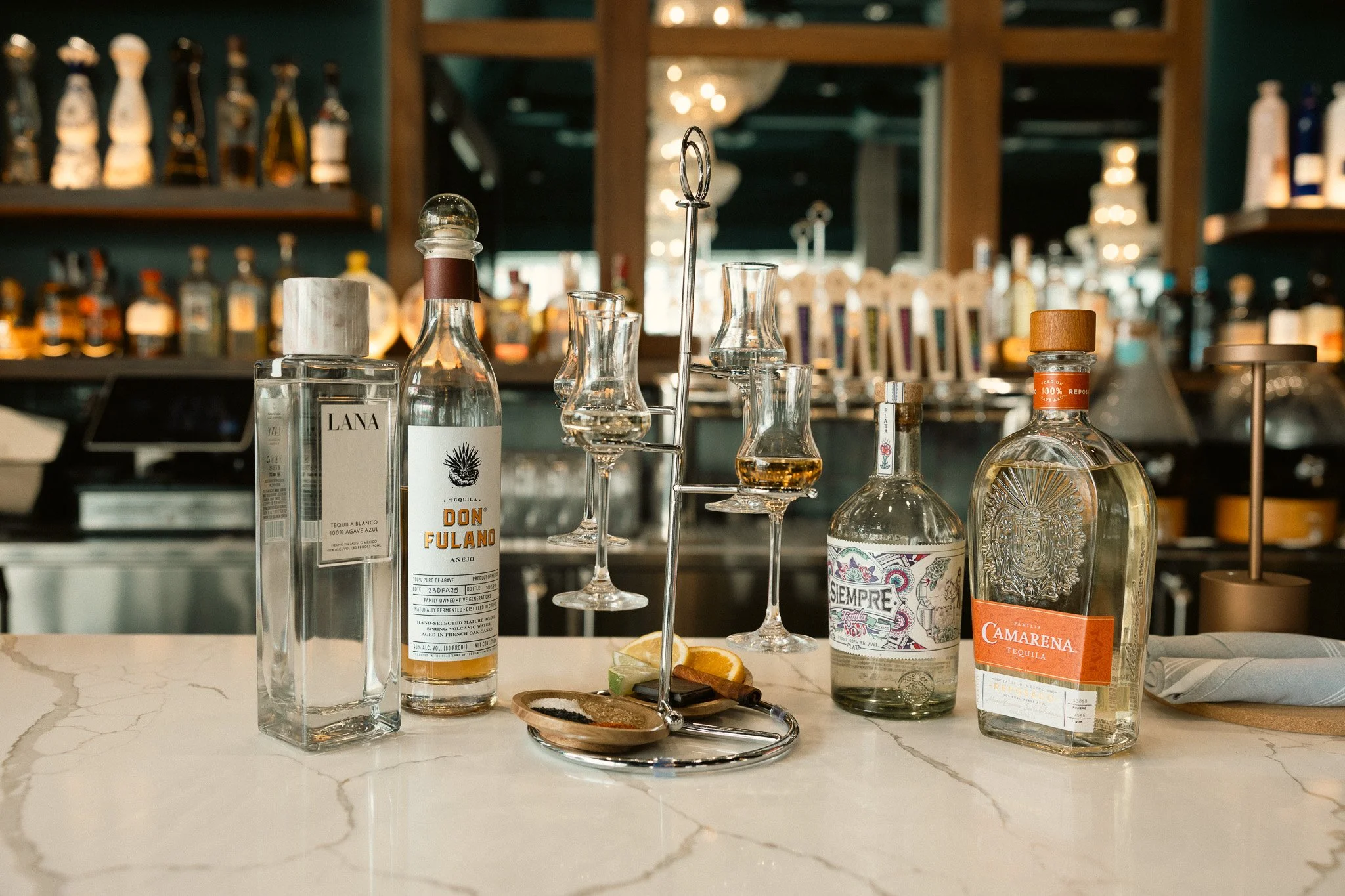 Bar counter with various liquor bottles and glassware, including tequila bottles, cocktail glasses, and a small wooden tray with salt and lemon slices, in a dimly lit bar or restaurant setting.