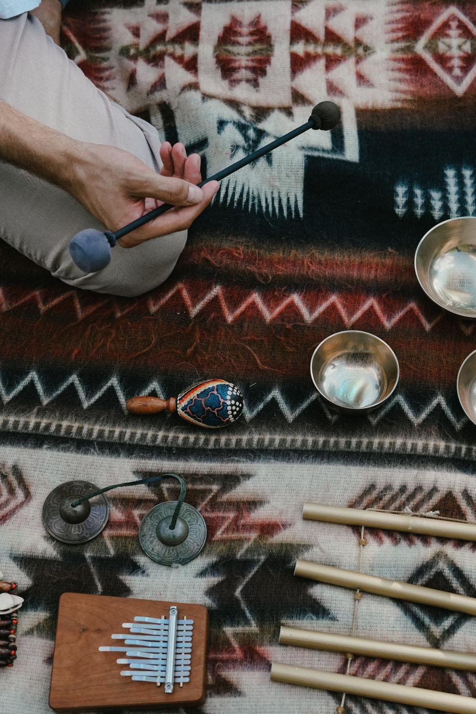 Person holding a mallet or hammer above metal singing bowls, on a patterned textile with various musical instruments including a rattle, chimes, a wind instrument, and cups around them.