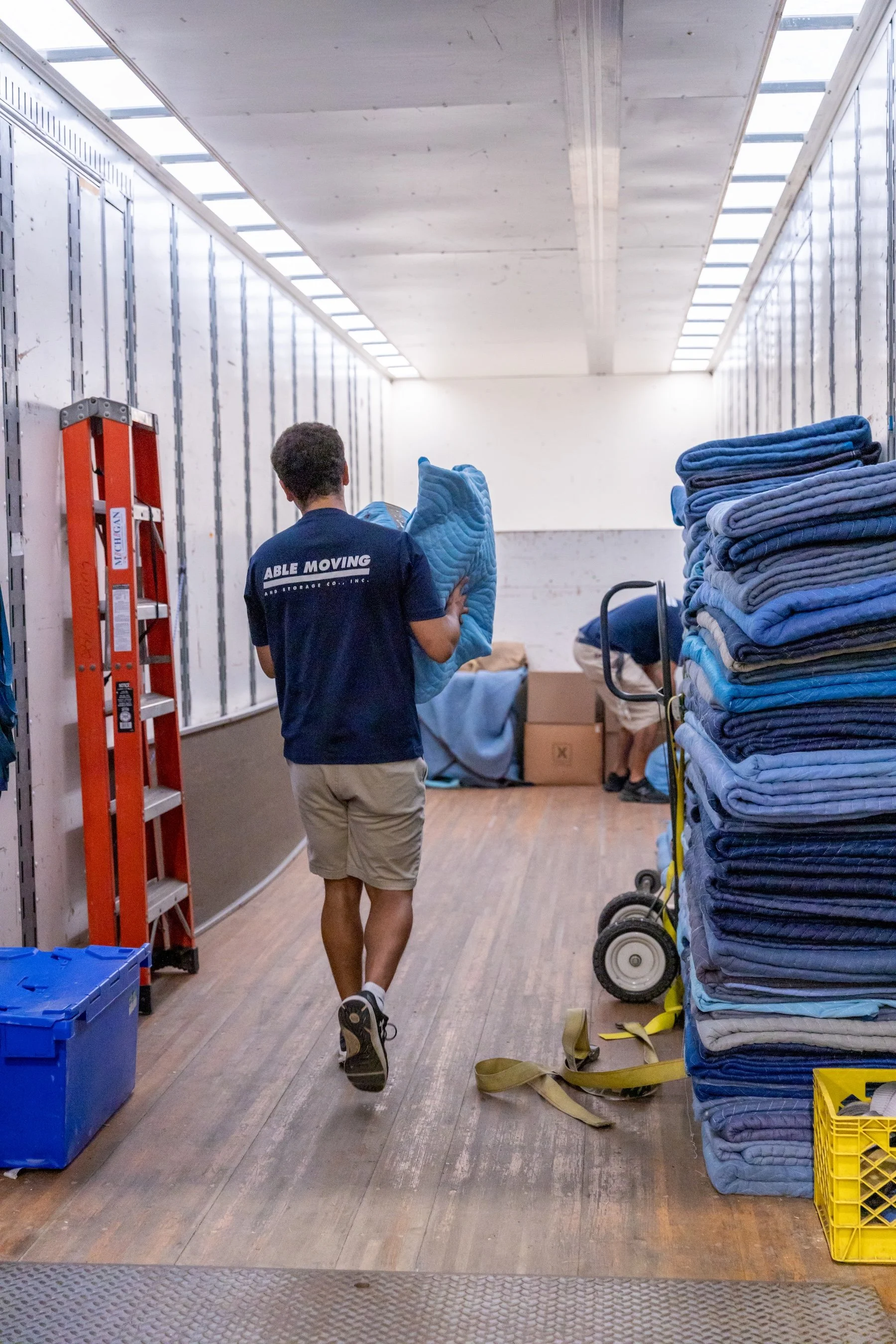 A man in shorts and a navy T-shirt walking through a storage room with stacked blue and gray clothing, a red ladder, and various boxes.