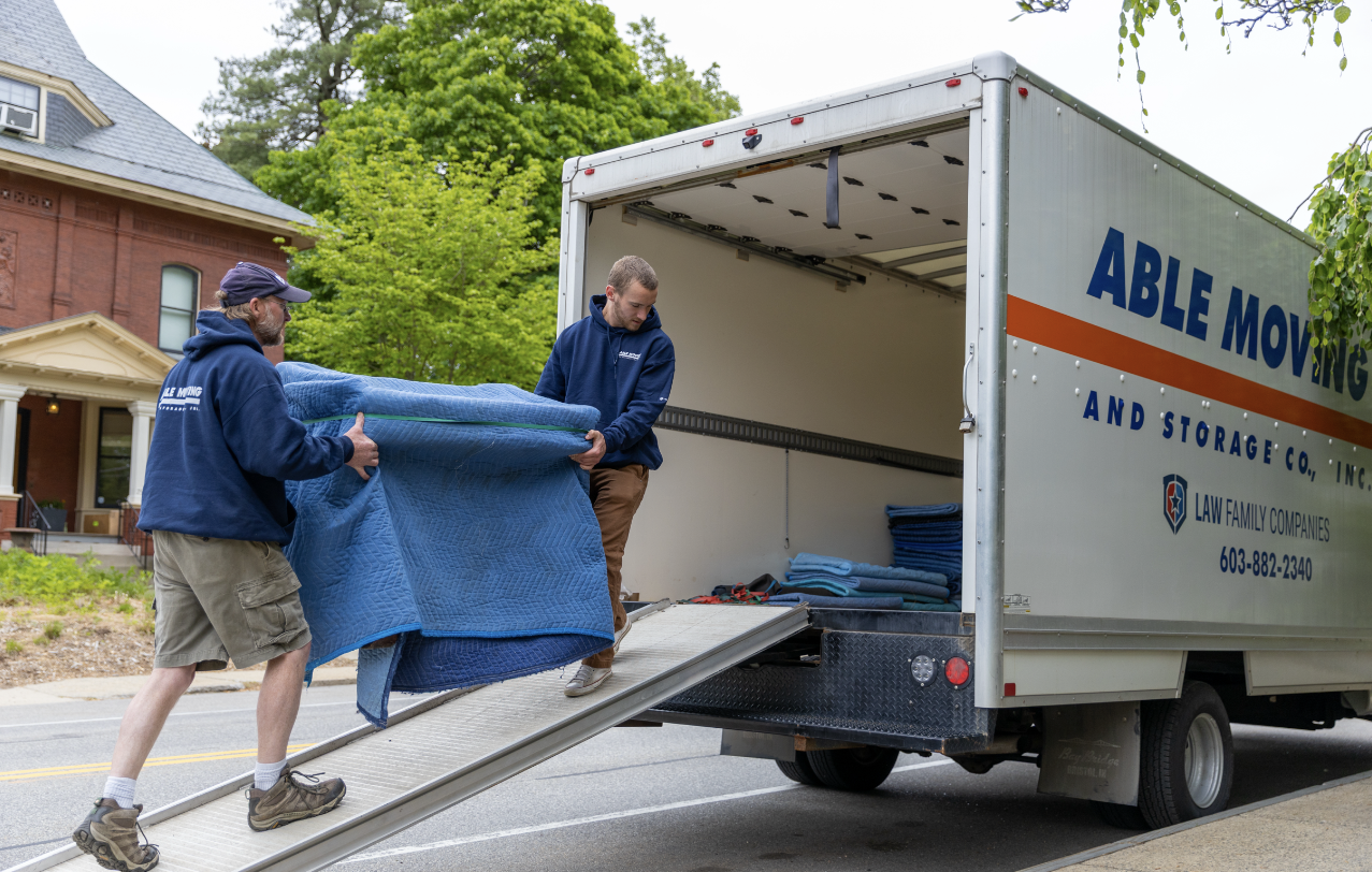 Two men loading a moving truck with blankets on a suburban street. One man is on the ramp holding a large blue blanket, and the other is inside the truck organizing additional blankets.
