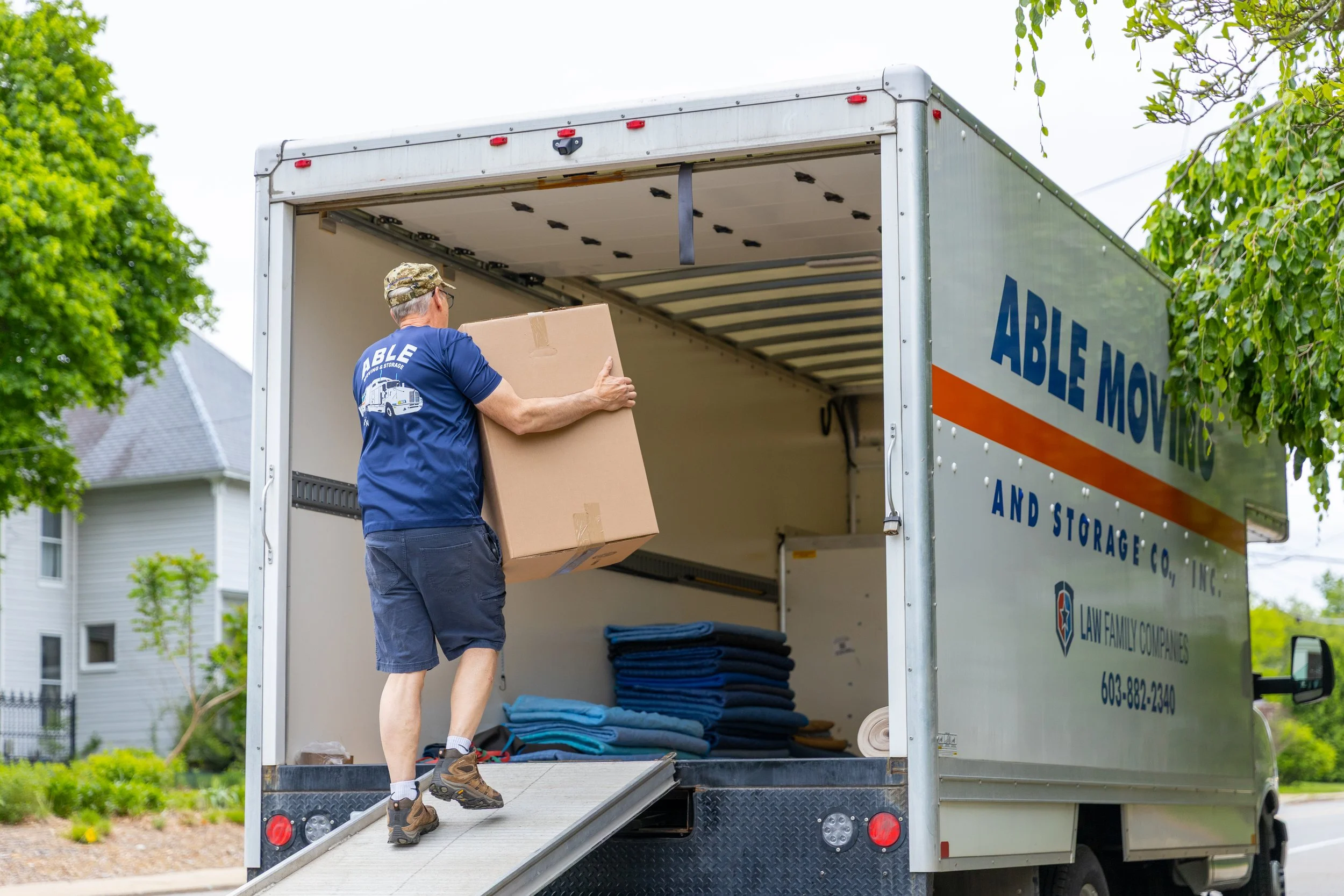 A man wearing a blue shirt and shorts is carrying a cardboard box into a moving truck with open back doors, parked outdoors near residential buildings and trees.