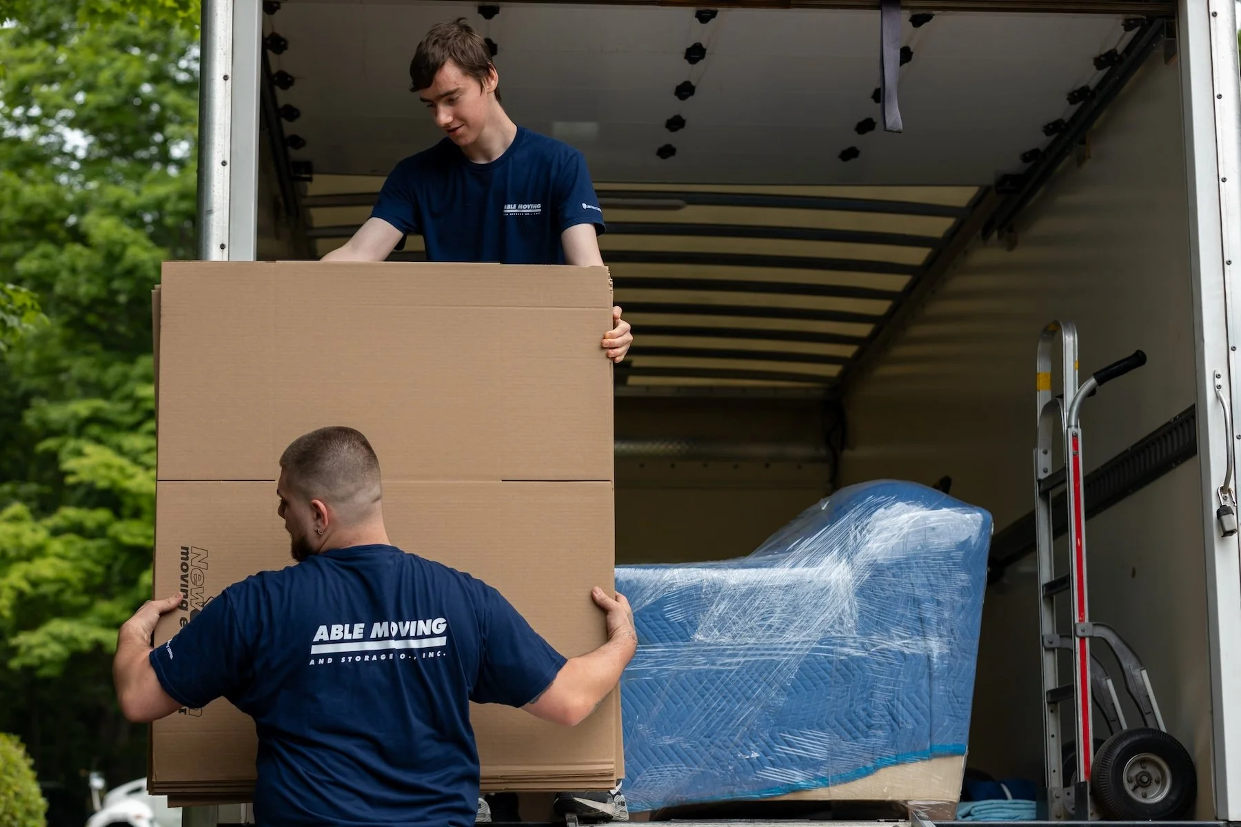 Two movers loading large boxes onto a truck, with one mover holding a big cardboard box and the other helping to steady it. The truck is open, and there are various wrapped items inside. The movers are wearing navy blue shirts with the company name "Able Moving" on the back.