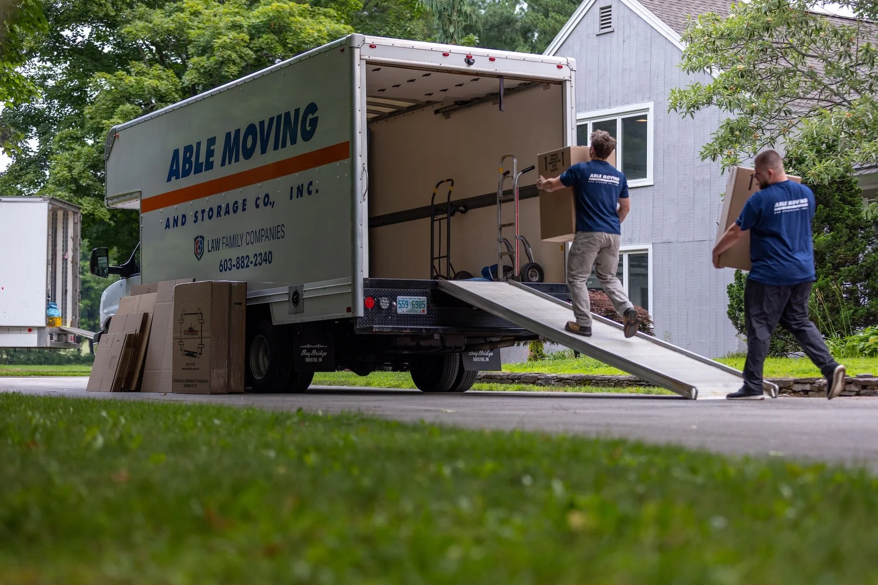 Two movers from Able Moving unload boxes from a moving truck parked in a residential driveway. One mover is carrying a box up a ramp, and the other is walking with a box. There are cardboard boxes on the ground and a house with gray siding in the background.