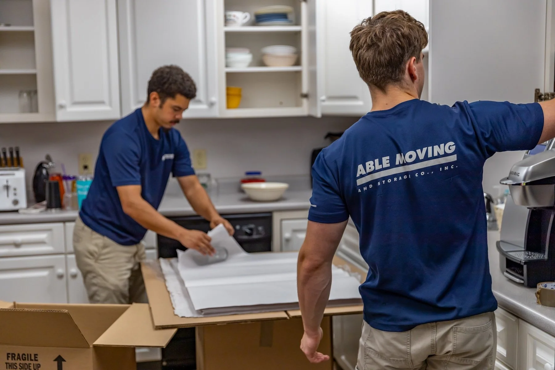 Two men in blue shirts working in a kitchen, packing or preparing items on a table for moving.