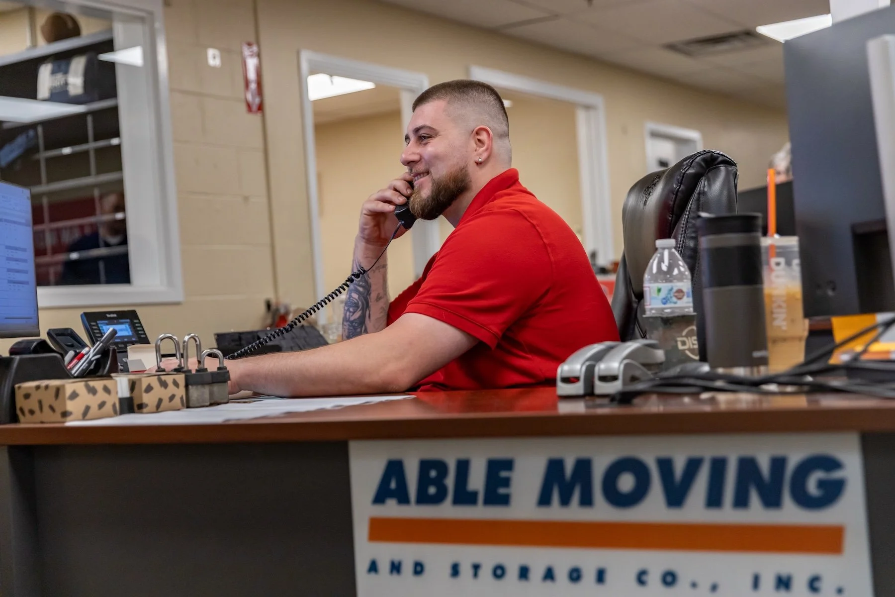 A smiling man with a beard and tattoos on his arm, wearing a red shirt, sitting at a desk and talking on a corded phone.
