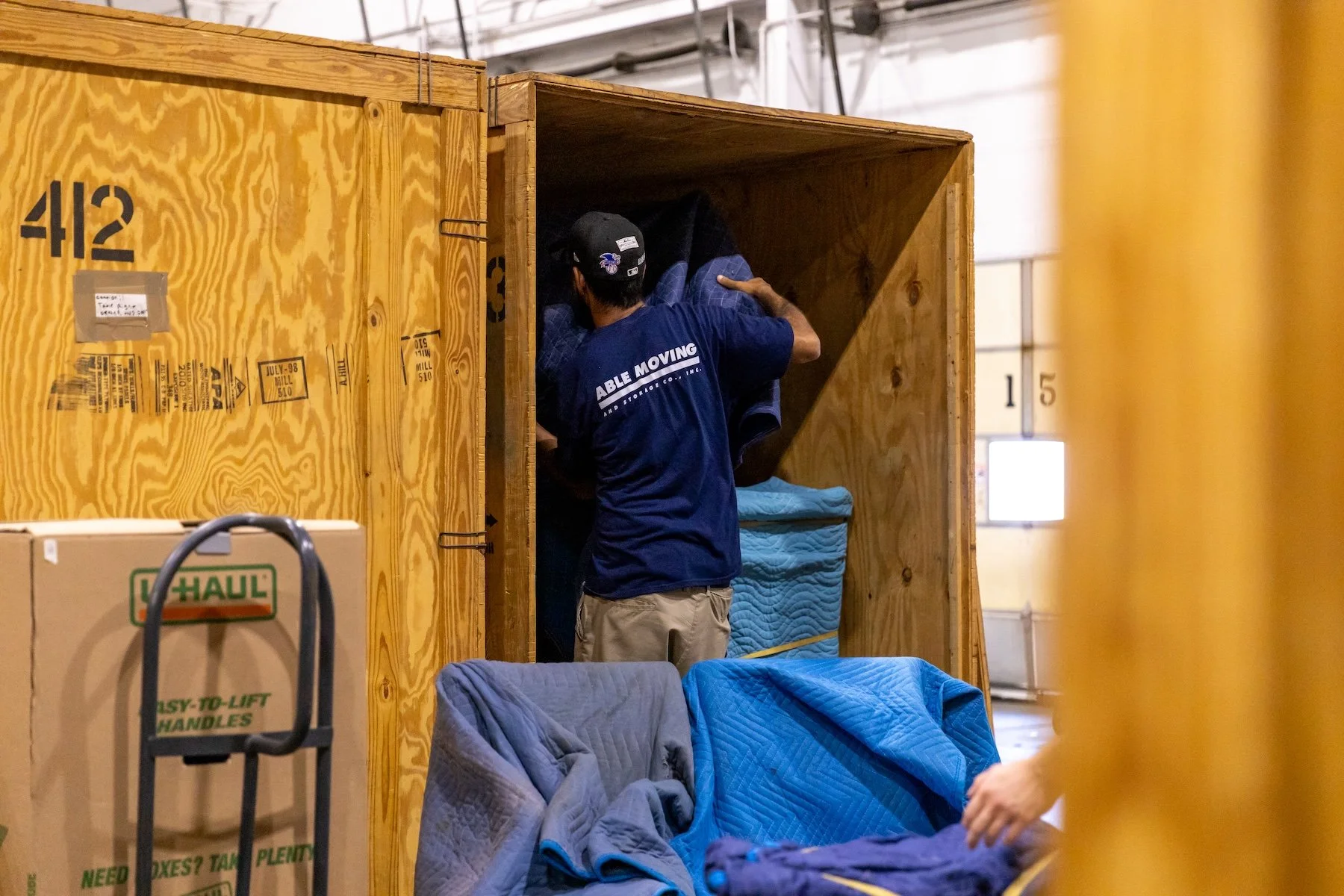 A man wearing a blue shirt and a baseball cap is packing items into a wooden storage unit. There are boxes and blankets in the area, indicating move-in activity.