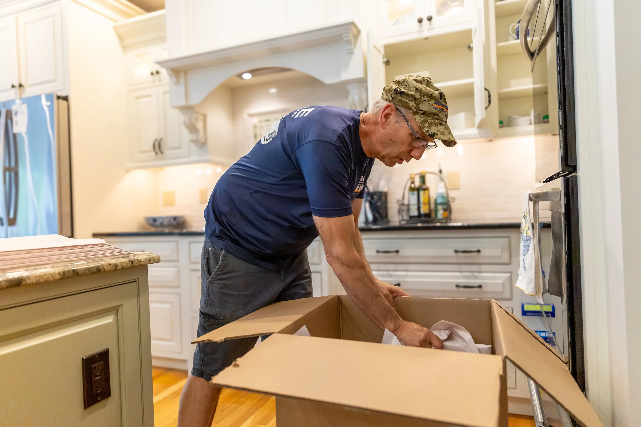 An older man with glasses and a camouflage cap opening a cardboard box in a kitchen with white cabinets and beige countertops.