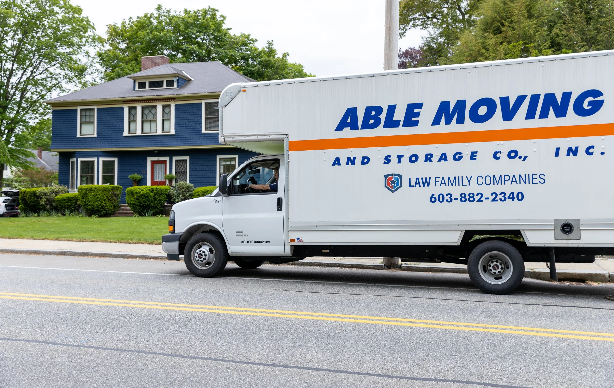 Moving truck on a street with a blue house in the background.