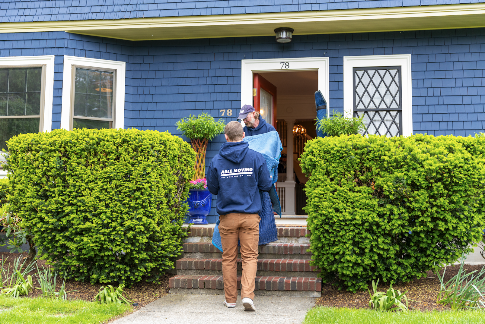 A moving crew unloading items at a blue house with the number 78 on the door. The crew member in the foreground is wearing a navy blue hoodie with "Able Moving" printed on the back, while another is passing a large wrapped item to a man at the door.