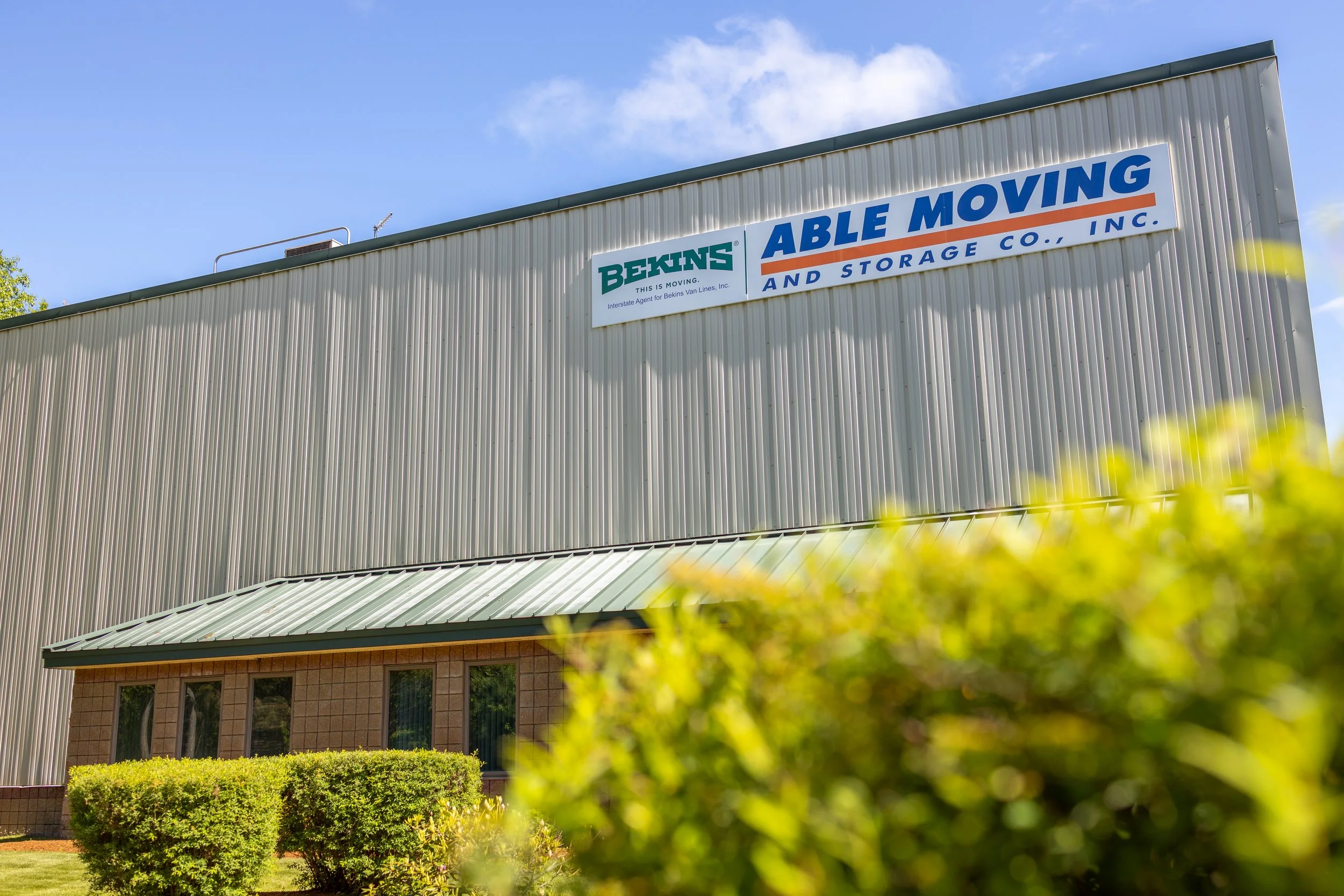 Sign on building reads 'Bekins Able Moving and Storage Inc.' with a green and blue logo, beige industrial-style building, blue sky, green bushes in foreground.