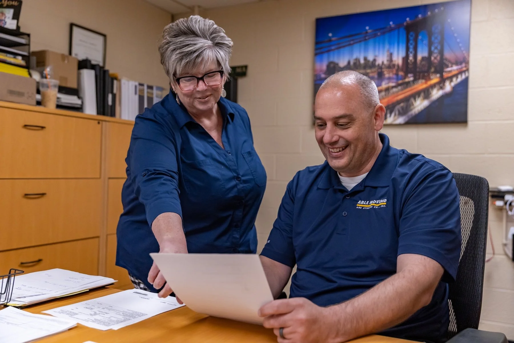 A woman and a man wearing matching blue shirts are smiling and looking at a document in an office, with papers on the desk and a large wall photo of a bridge in the background.