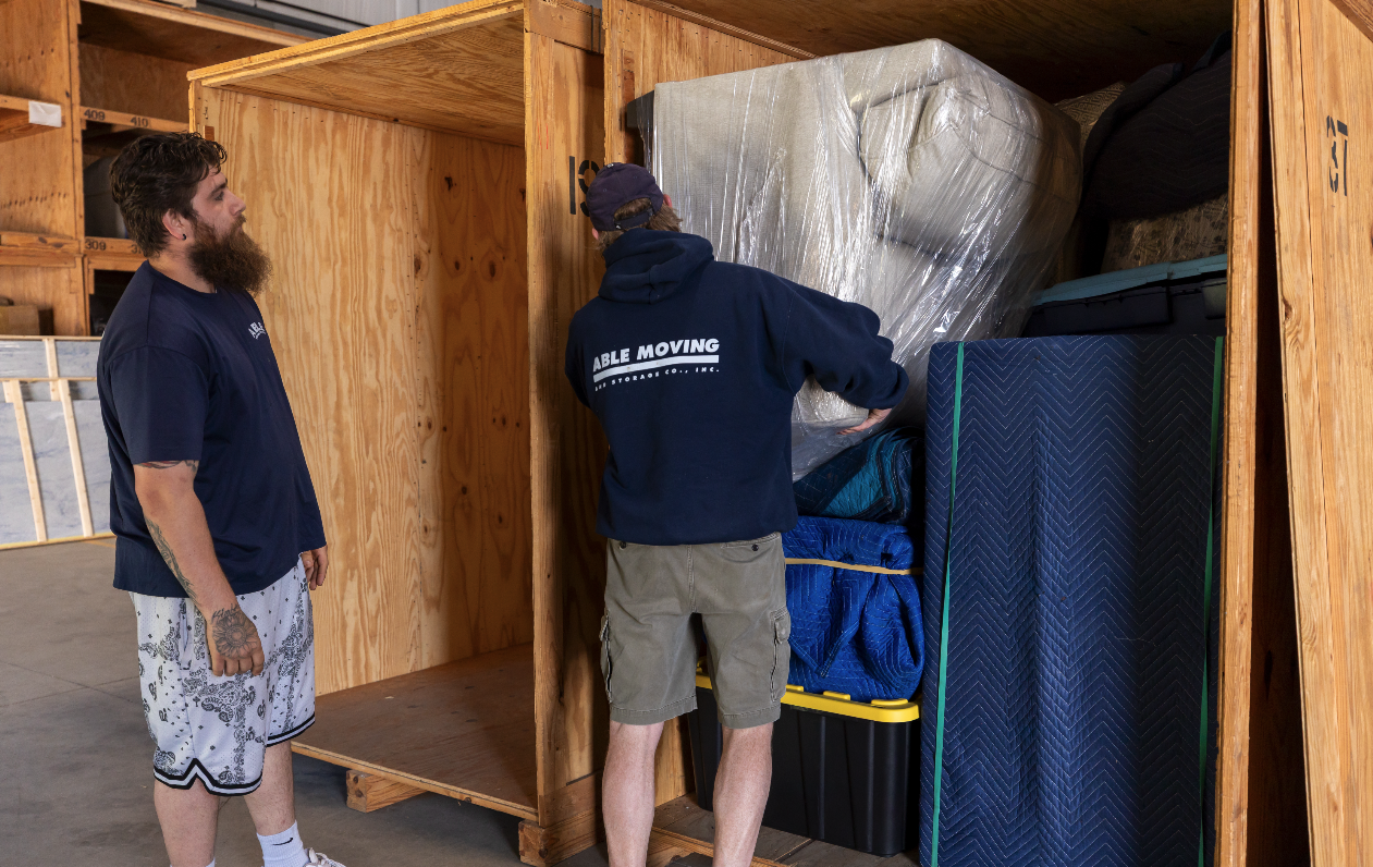 Two men packing or organizing items into a wooden storage crate in a warehouse. One man is facing inside the crate, and the other is watching.