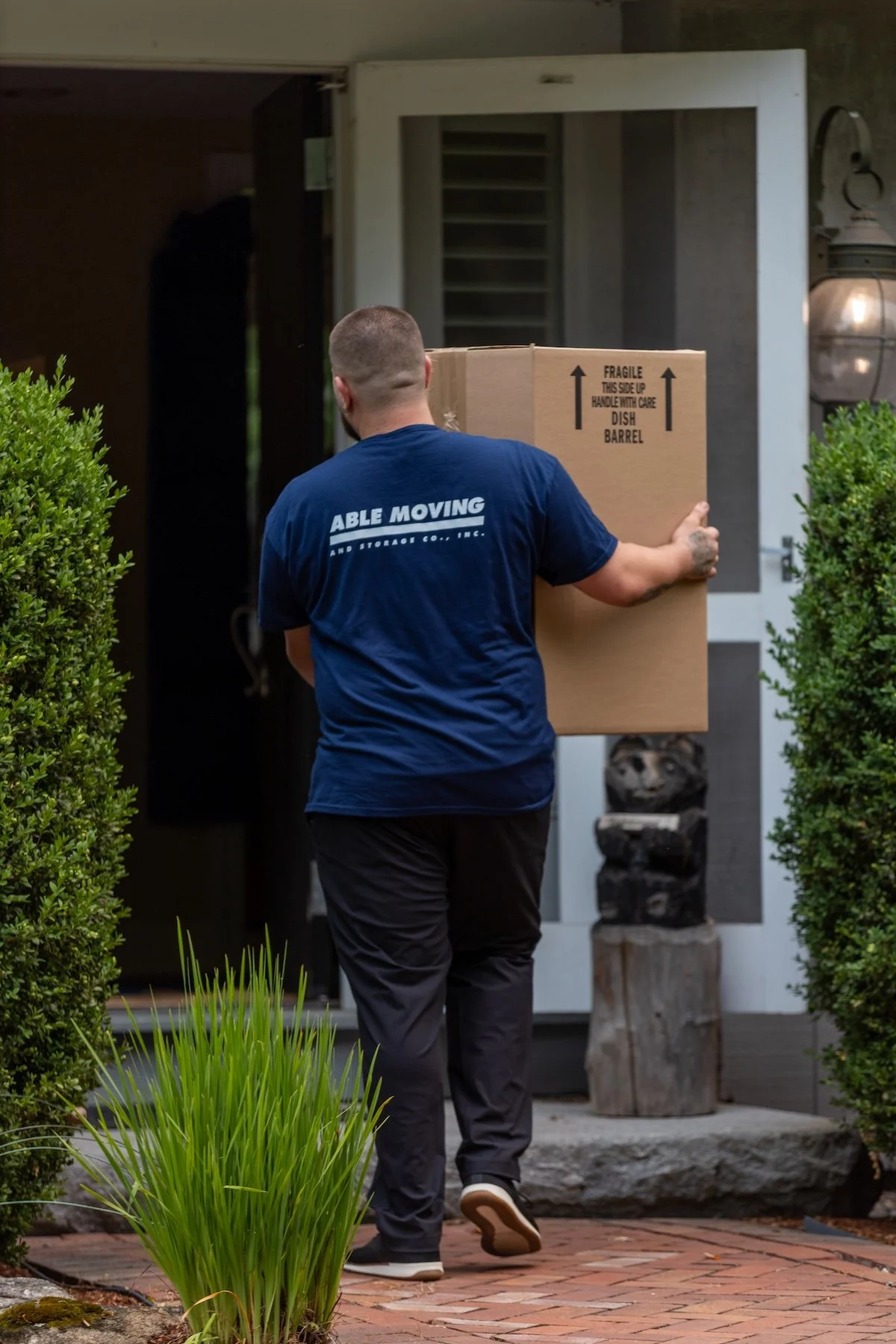 A man carrying a large cardboard box labeled 'Fragile, this side up, handle with care, dish barrel' walking towards a house entrance. The man is wearing a blue T-shirt with the text "Able Moving and Storage Co., Inc." and dark pants. The house has bushes on either side of the walkway, and there is a decorative log and planter on the front porch.