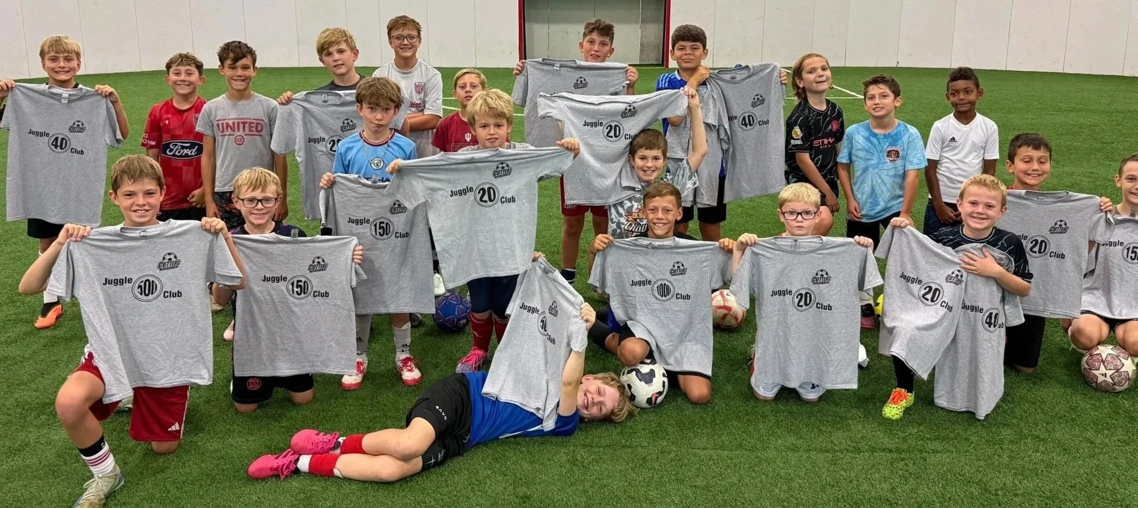 A group of young boys and girls on a soccer field, holding up gray T-shirts with the text 'Juggle 20 Club' and numbers, some kneeling and some standing, with soccer balls in front and around them.