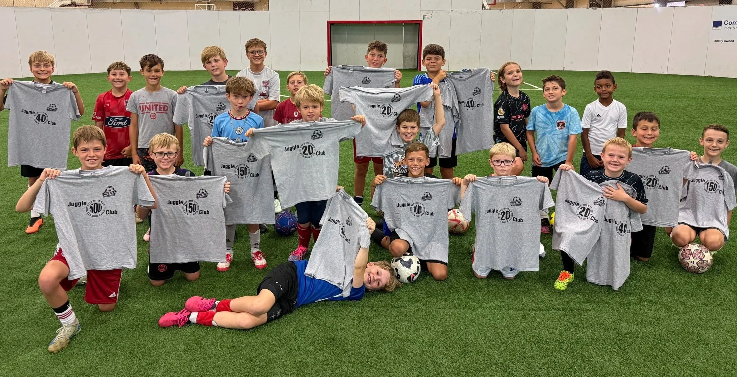 A group of children on a soccer field holding up gray T-shirts with various numbers and text, smiling at the camera after a game or practice.