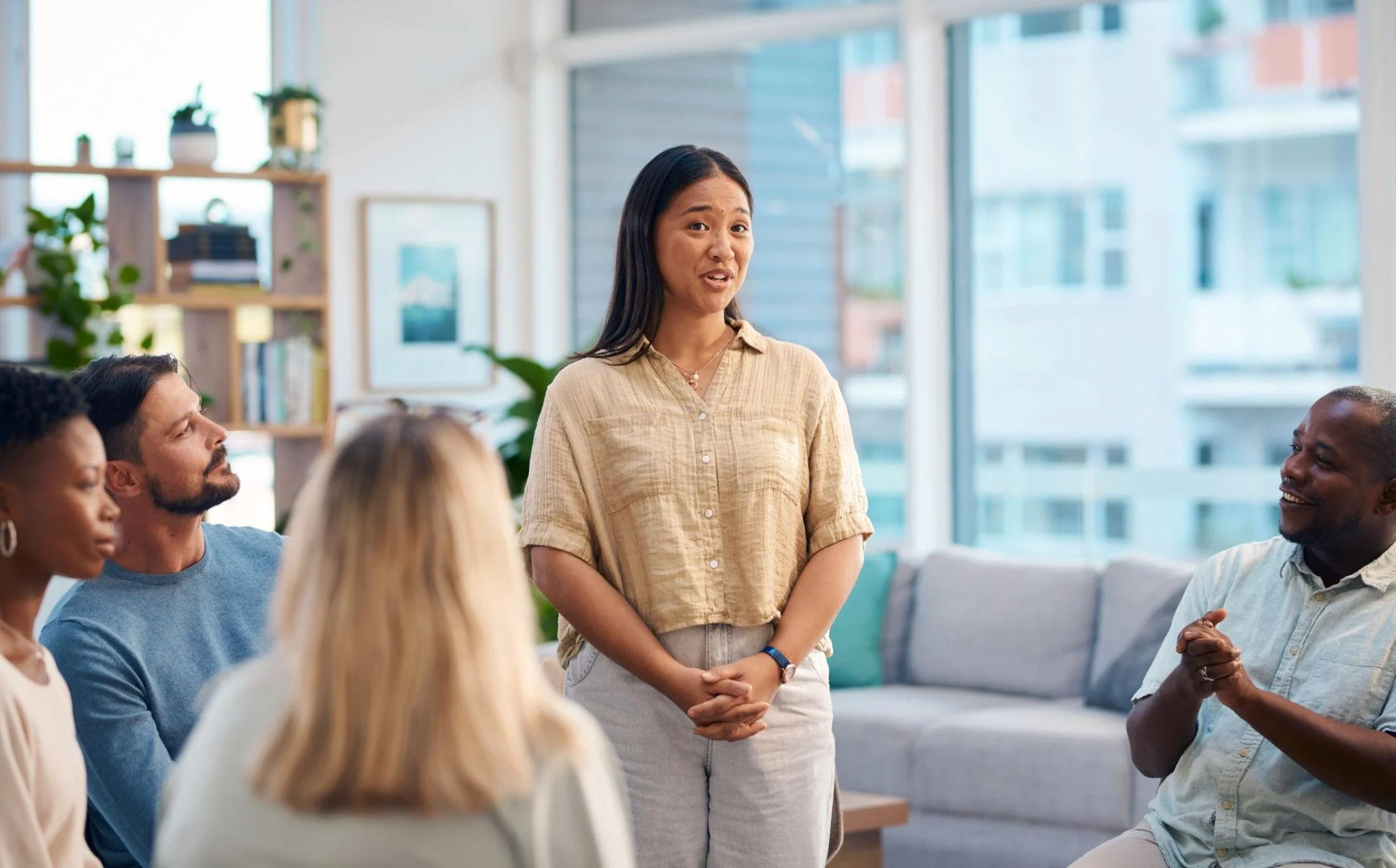 A woman standing and speaking to a group of seated people in a bright office with large windows and plants.