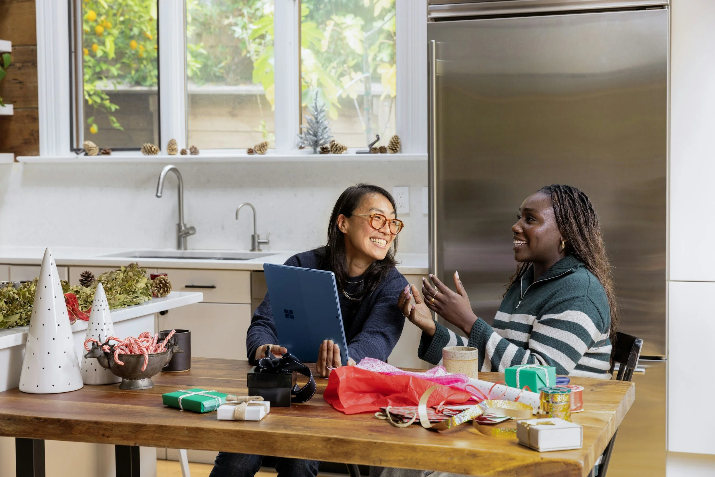 Two women sitting at a kitchen table, laughing and talking, surrounded by holiday gift wrapping and decorations, with a window showing greenery outside.