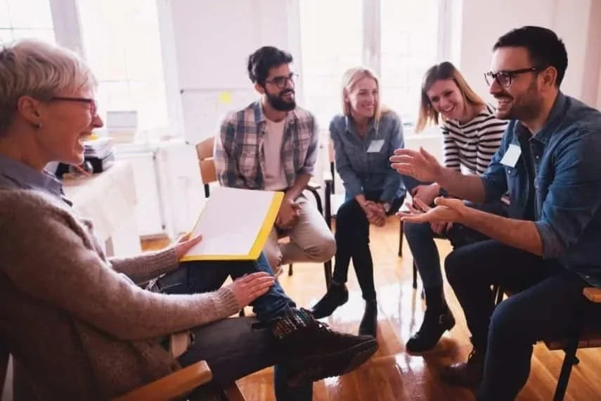 Group of five people sitting in a circle during a discussion or meeting in a bright room, smiling and engaging with each other.