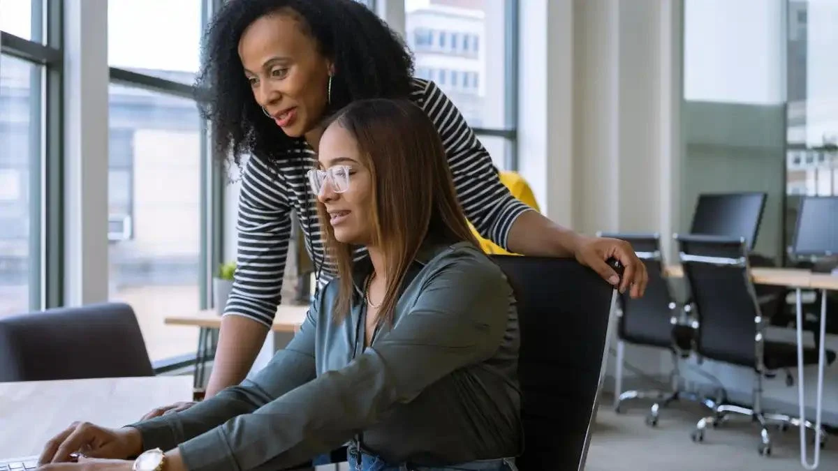 Two women in an office looking at a laptop together, one standing and leaning over the seated woman.