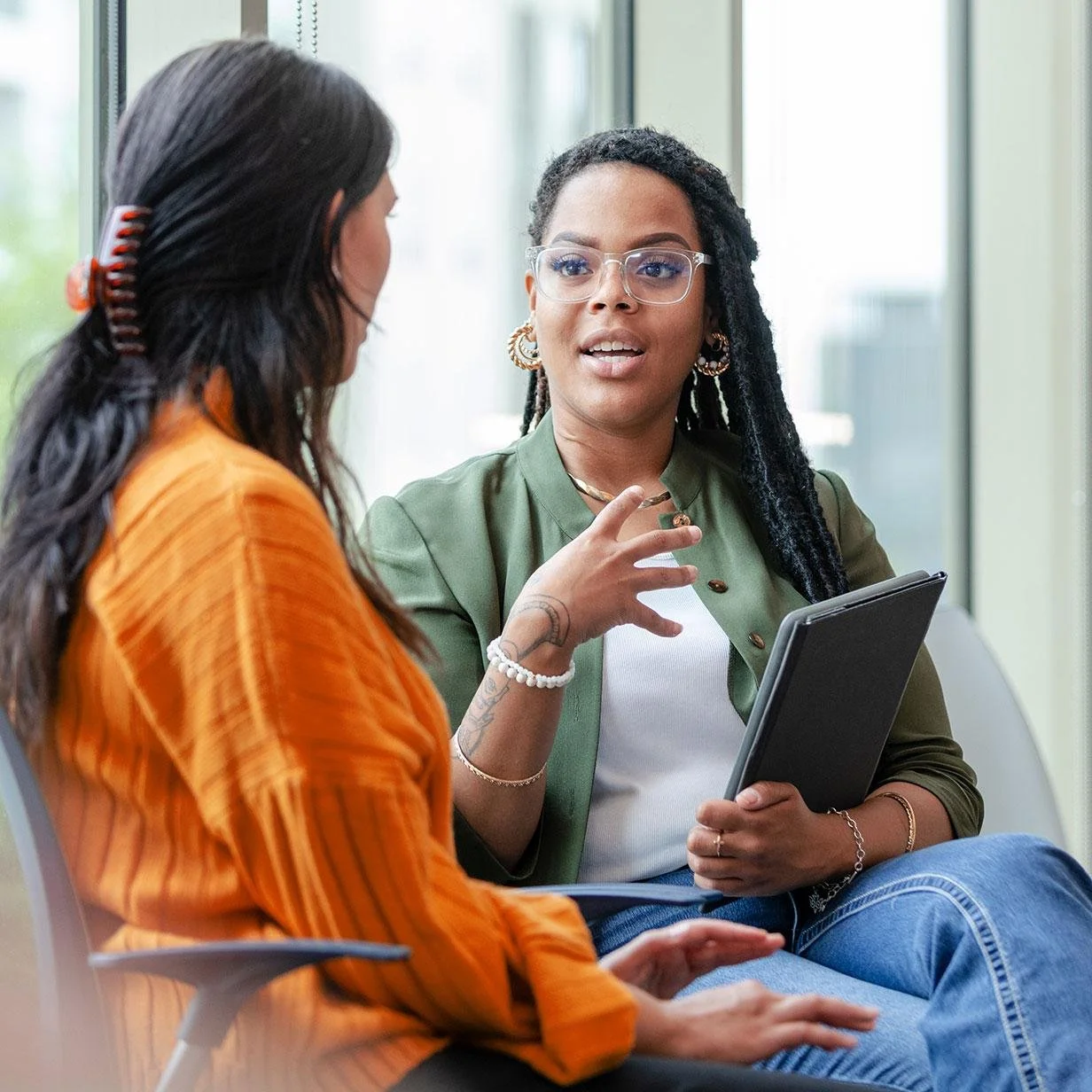 Two women having a conversation in an office with large windows, one holding a tablet and wearing glasses and jewelry, and the other with a hair clip and orange shirt.