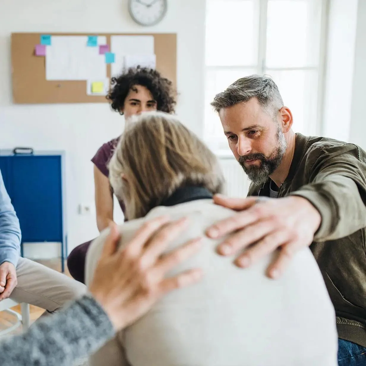 Group of people providing emotional support to an elderly woman in a bright room with a wall clock and bulletin board.