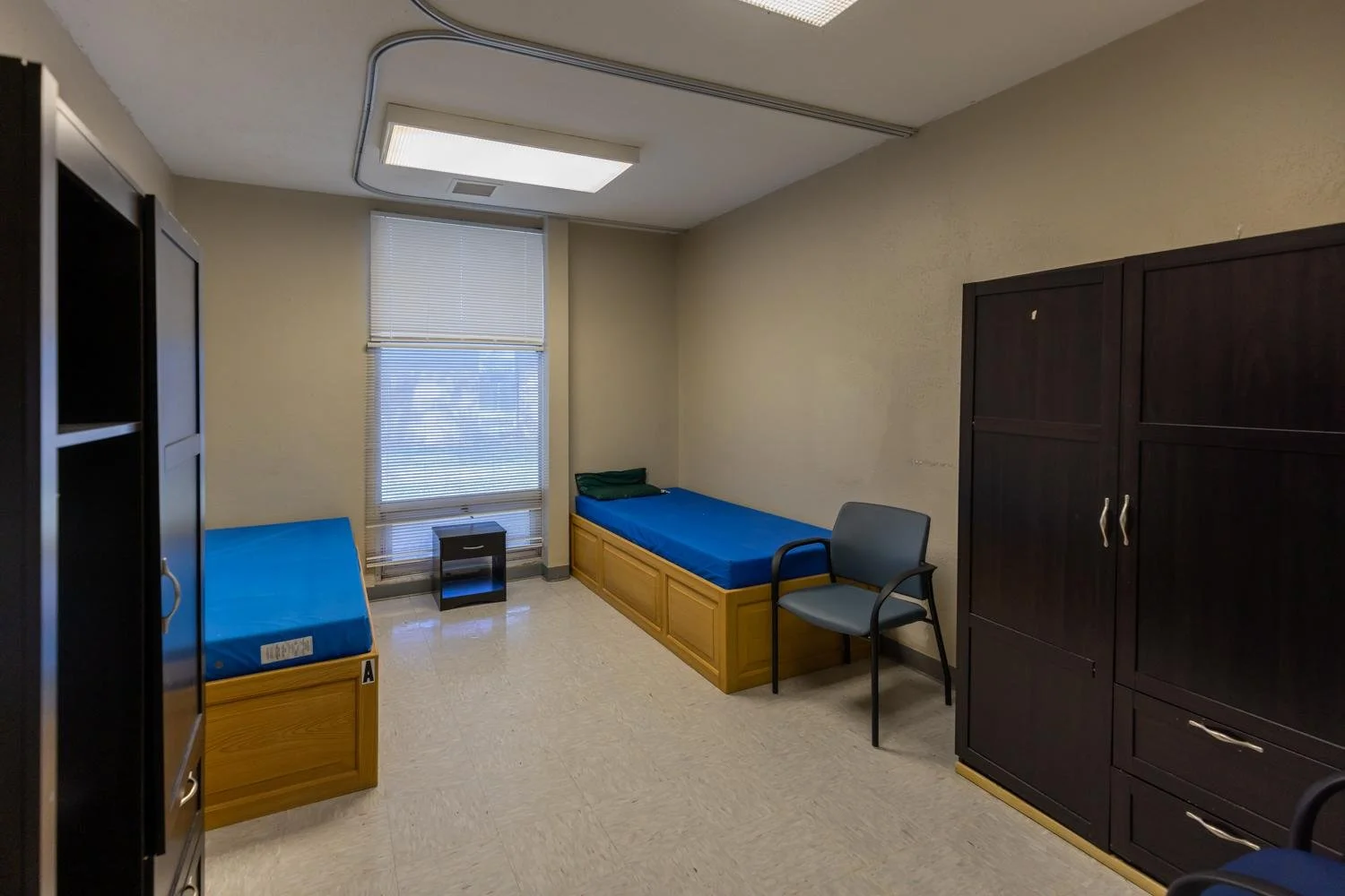 A small, sparsely furnished room with two single beds with blue mattresses, a black bedside table, a dark brown cabinet, a gray chair, and a window with blinds.