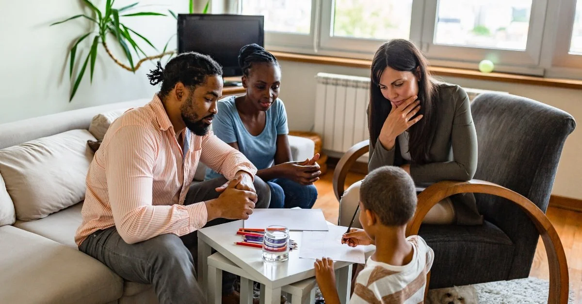 A diverse family gathered in a living room, talking around a small white table with drawing supplies, while a woman looks on thoughtfully and a young boy draws.