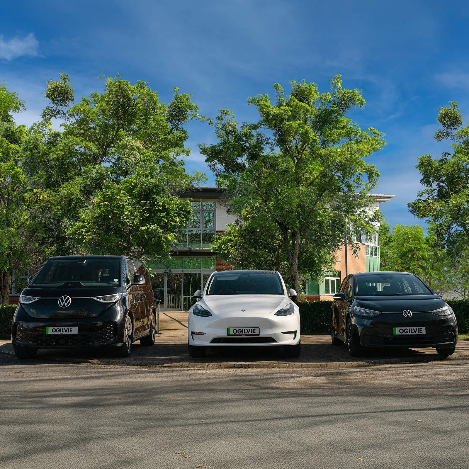 Three cars parked in front of a building with trees and a blue sky in the background. The cars have custom license plates reading "OGILVIE."