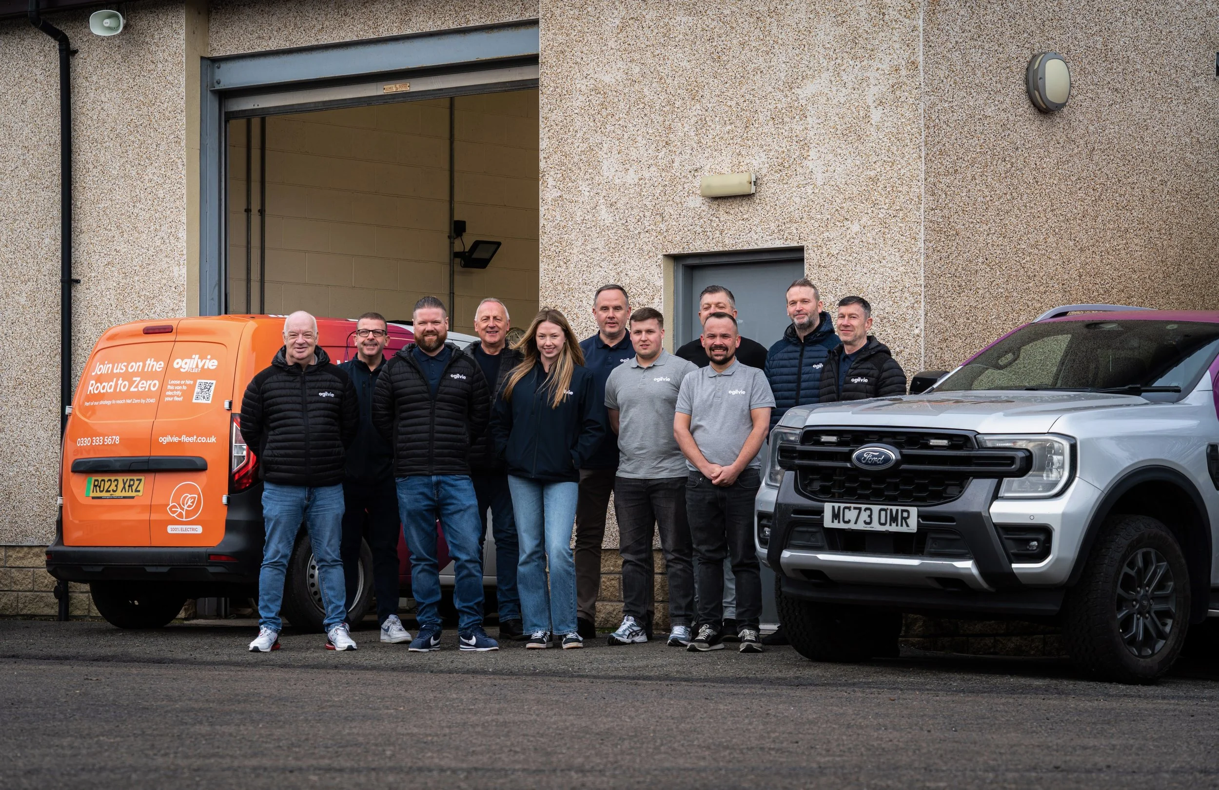 Group of eleven people standing outside near parked vehicles against a textured beige wall, including a silver Ford truck, an orange electric van with eco-friendly signage, and a maroon and silver van. The group appears to be a team or colleagues posing for a photo.