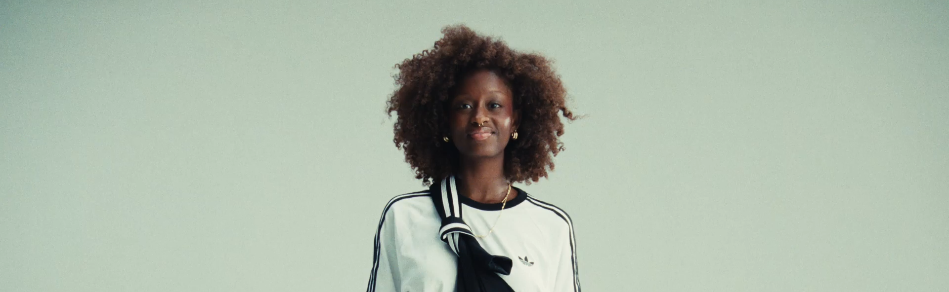 Een vrouw met een sprookjesachtige boskrullend haar, draag een witte top met zwarte strepen en een zwarte sweatshirt, en staat voor een effen lichtgroene achtergrond.