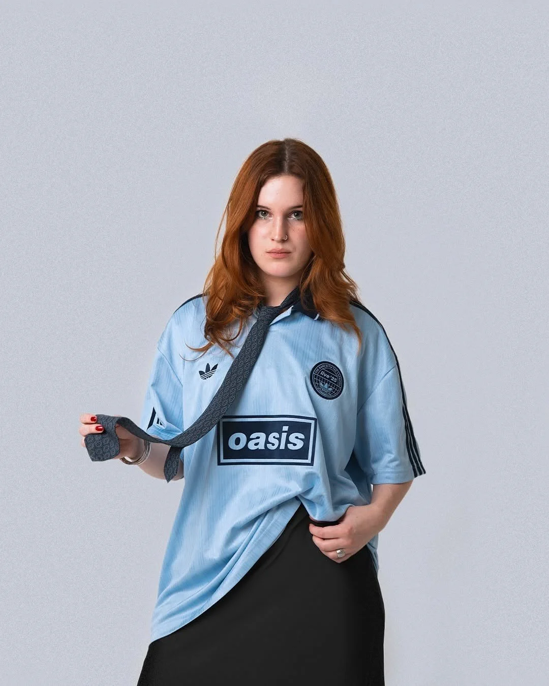 Vrouw met rood haar in een lichtblauw sportshirt met oasissponsor en Adidas-merken, die een touw vasthoudt, tegen een effen grijze achtergrond.