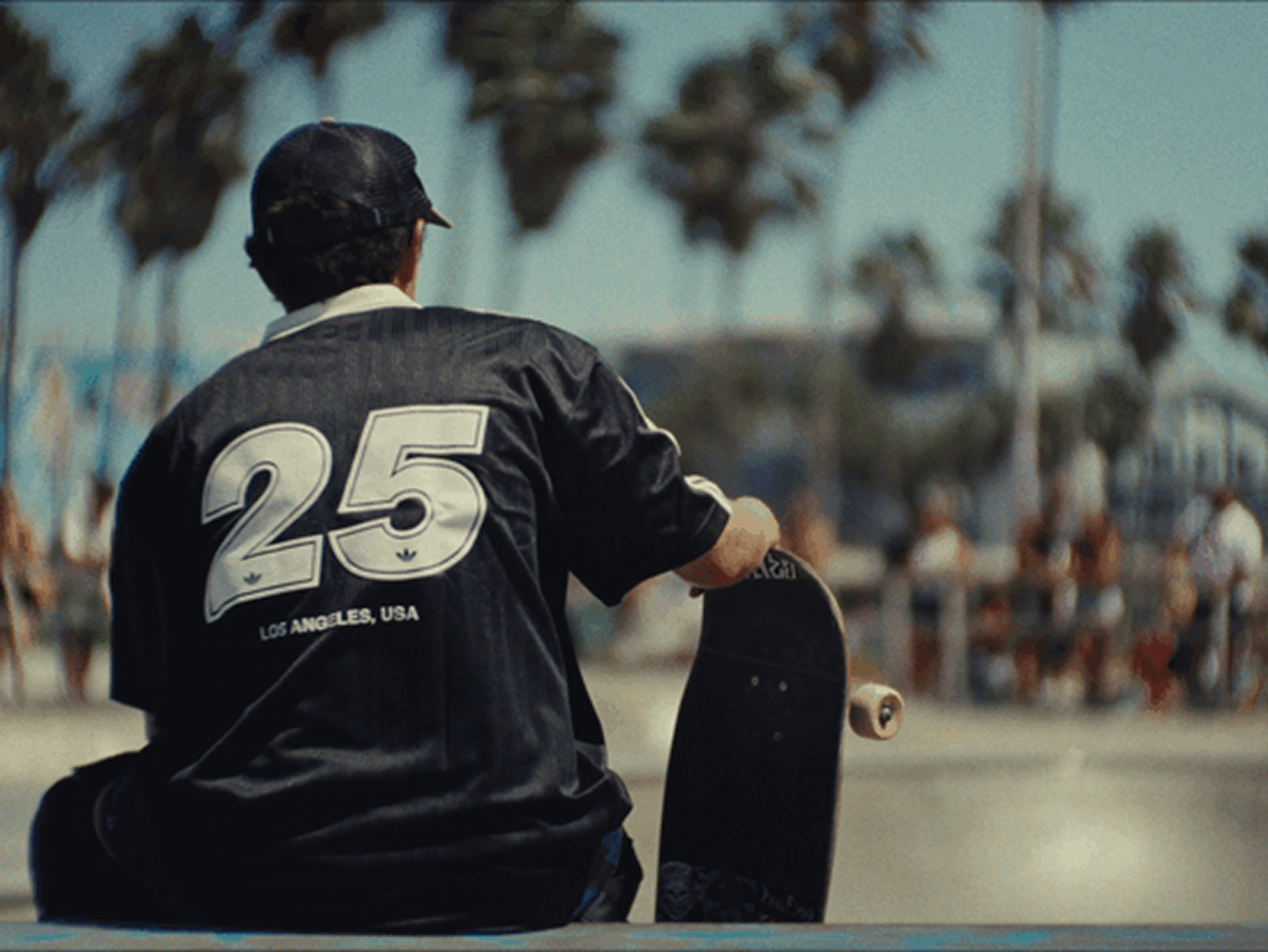 Een persoon die met een skateboard in een skatepark of buitenruimte staat. De persoon draagt een zwart jack met het nummer 25 en de tekst 'LOS ANGELES, USA' erop, en een zwarte pet.