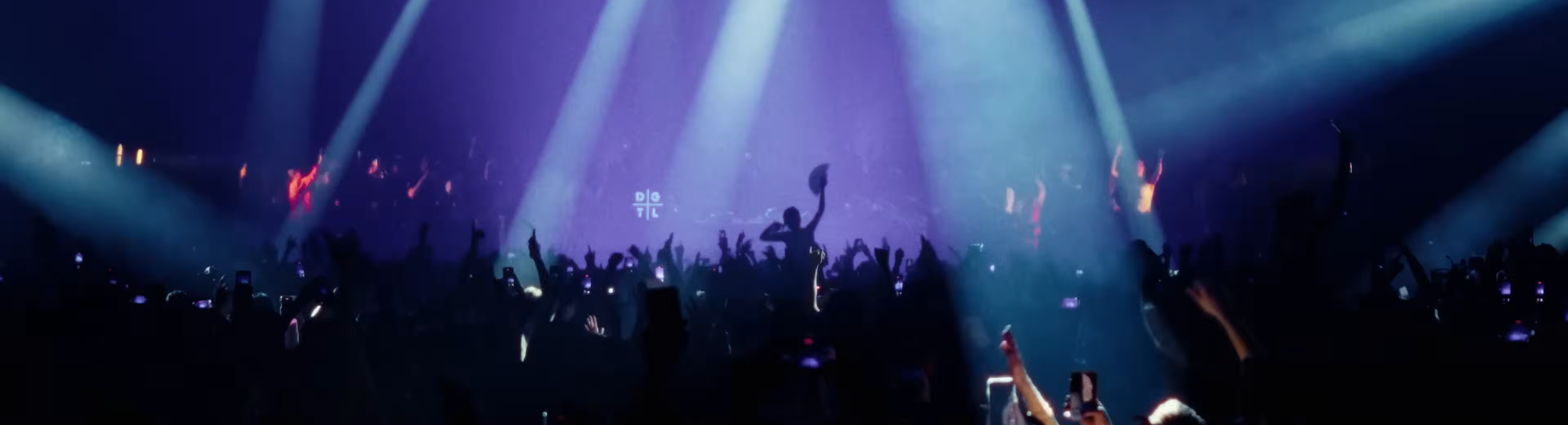 Een concert met een dansend publiek en een dj op het podium, verlicht door blauwe en paarse lichtstralen.