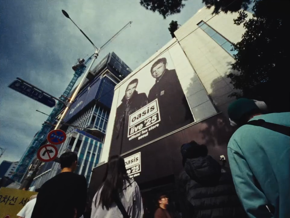 Straatzicht met mensen, een bouwlocatie met een hijskraan, en een groot billboard met een foto van twee mannen en teksten over Oasis Live 25.