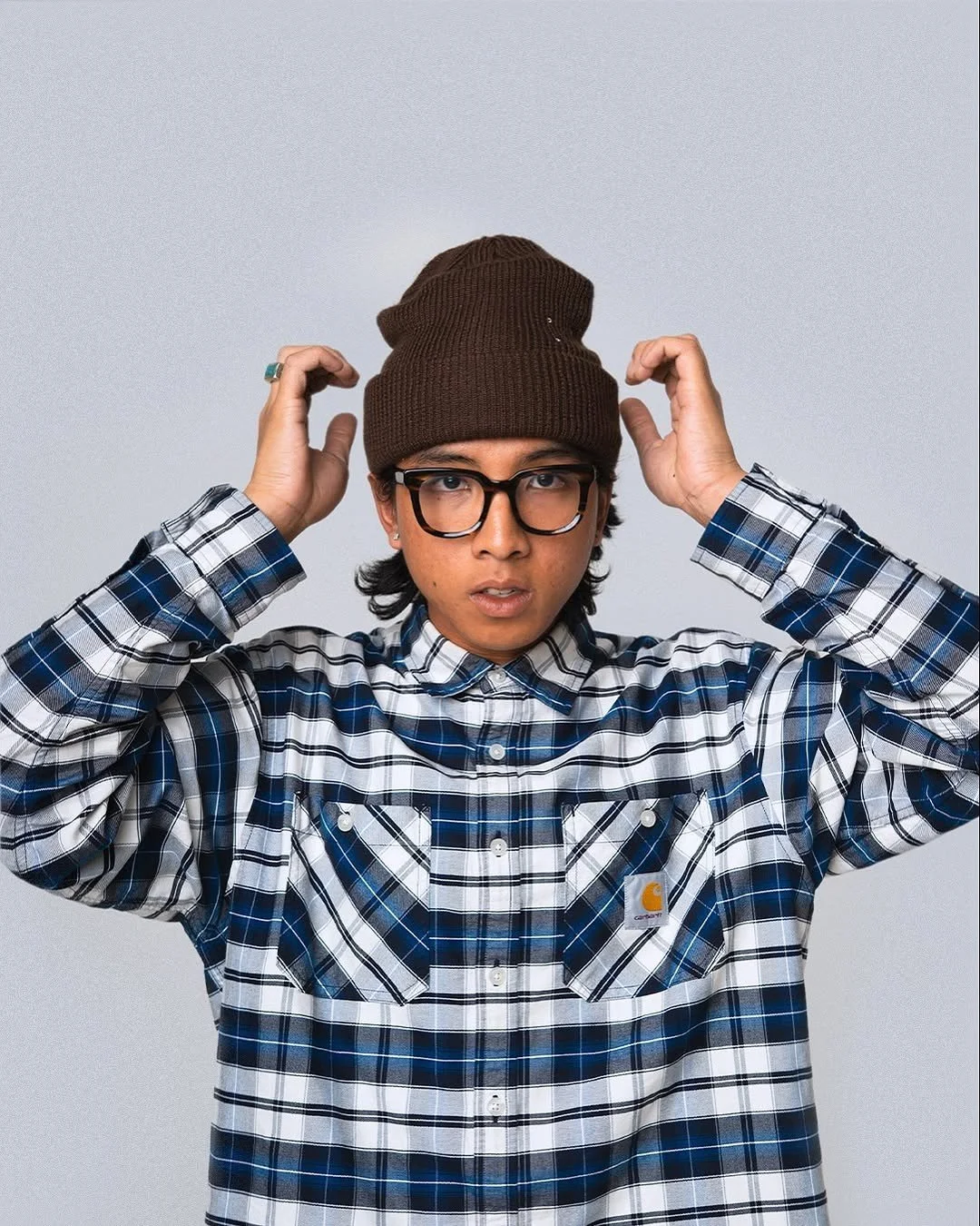 Een man met een bruin muts, zwarte bril, vlakke neus en zwart haar, draagt een blauw-wit geruite Carhartt jas en houdt zijn handen bij zijn hoofd.