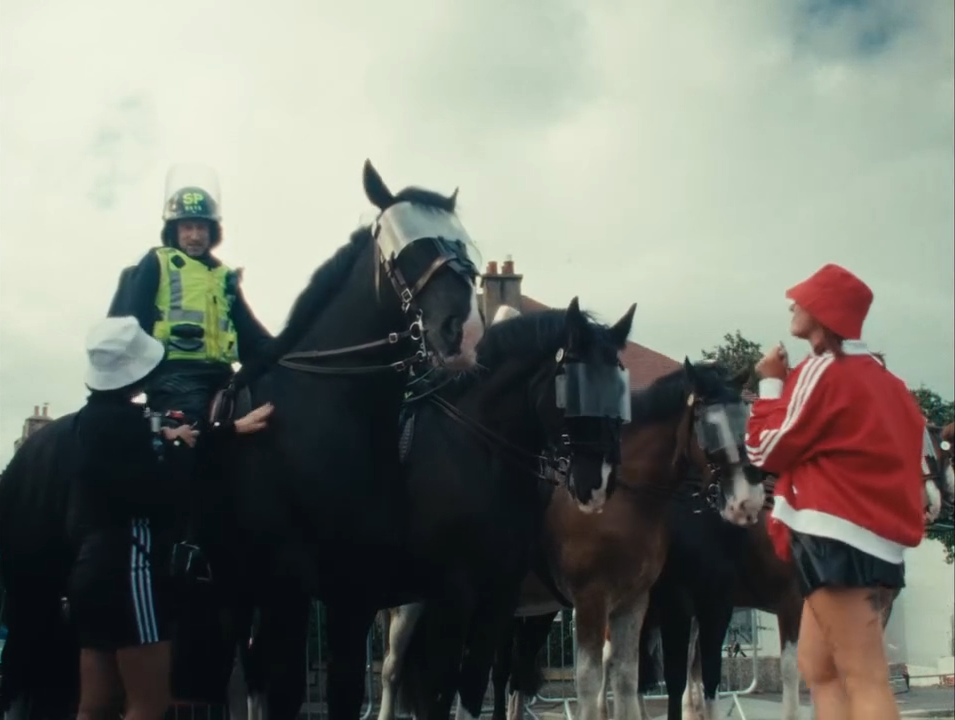 Politie op paarden praten met een vrouw in een rode jas en een rood mutsje in een openbare ruimte met huizen op de achtergrond.