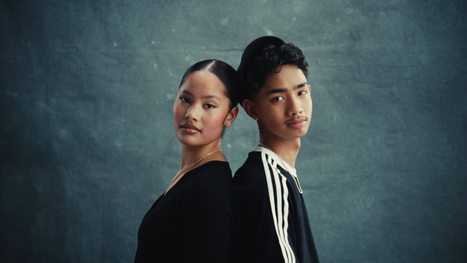 Een jonge vrouw en een jonge man staan met hun rug tegen elkaar, zij kijken naar de camera. Ze dragen moderne kleding en hebben kalme uitdrukkingen.