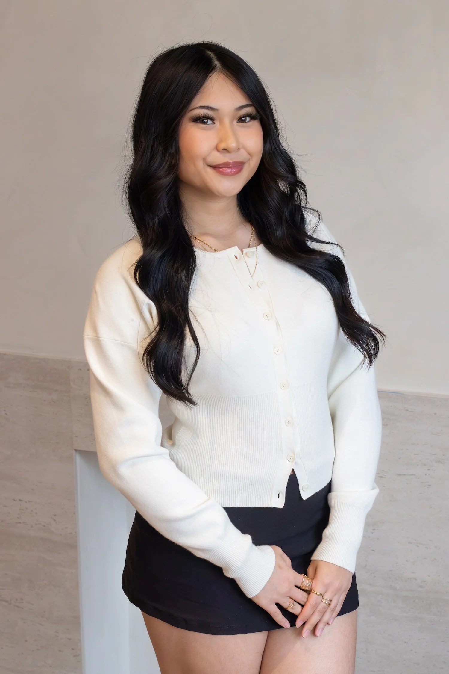 A beautiful young filipino woman stands confidently in front of a beige background wearing a white long sleeve top.