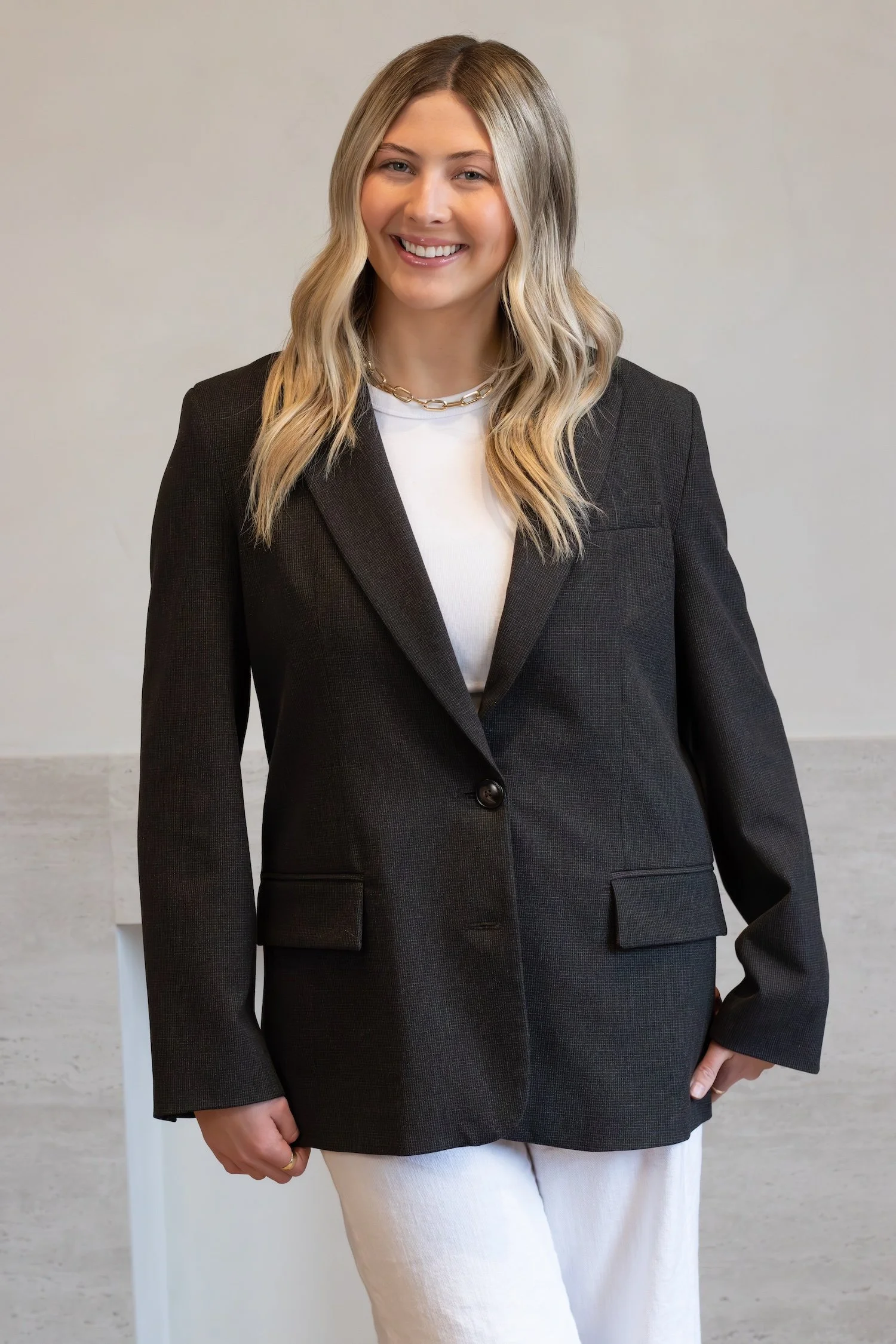 A beautiful young blonde girl smiles brightly as she wears a black blazer in front of a beige background.
