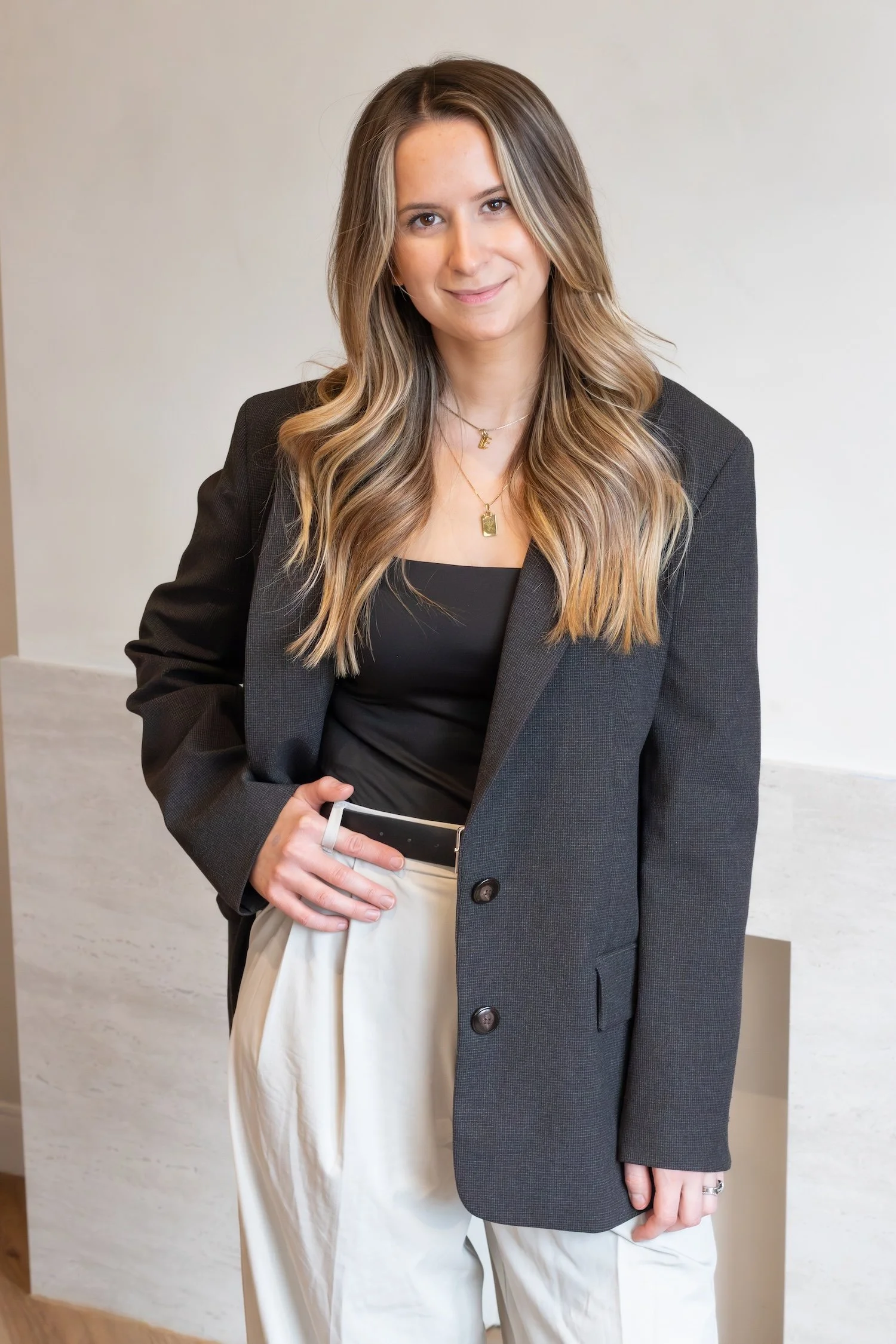 A beautiful, young, brown haired girl stands confidently in front of a beige backdrop as she wears a grey women's blazer.