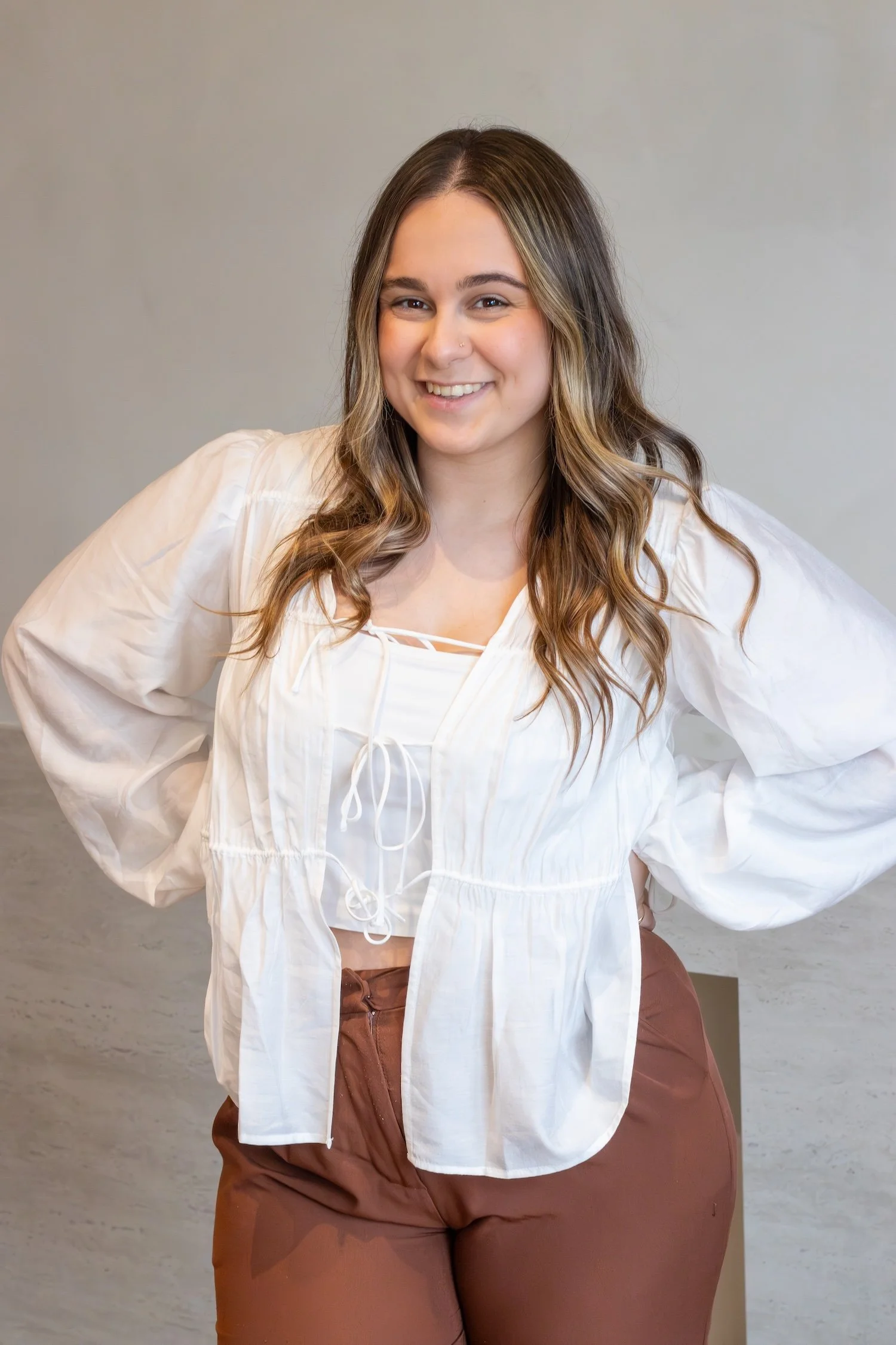 A beautiful brown haired girl stands smiling with her hands at her sides wearing a white lace top in front of a beige background