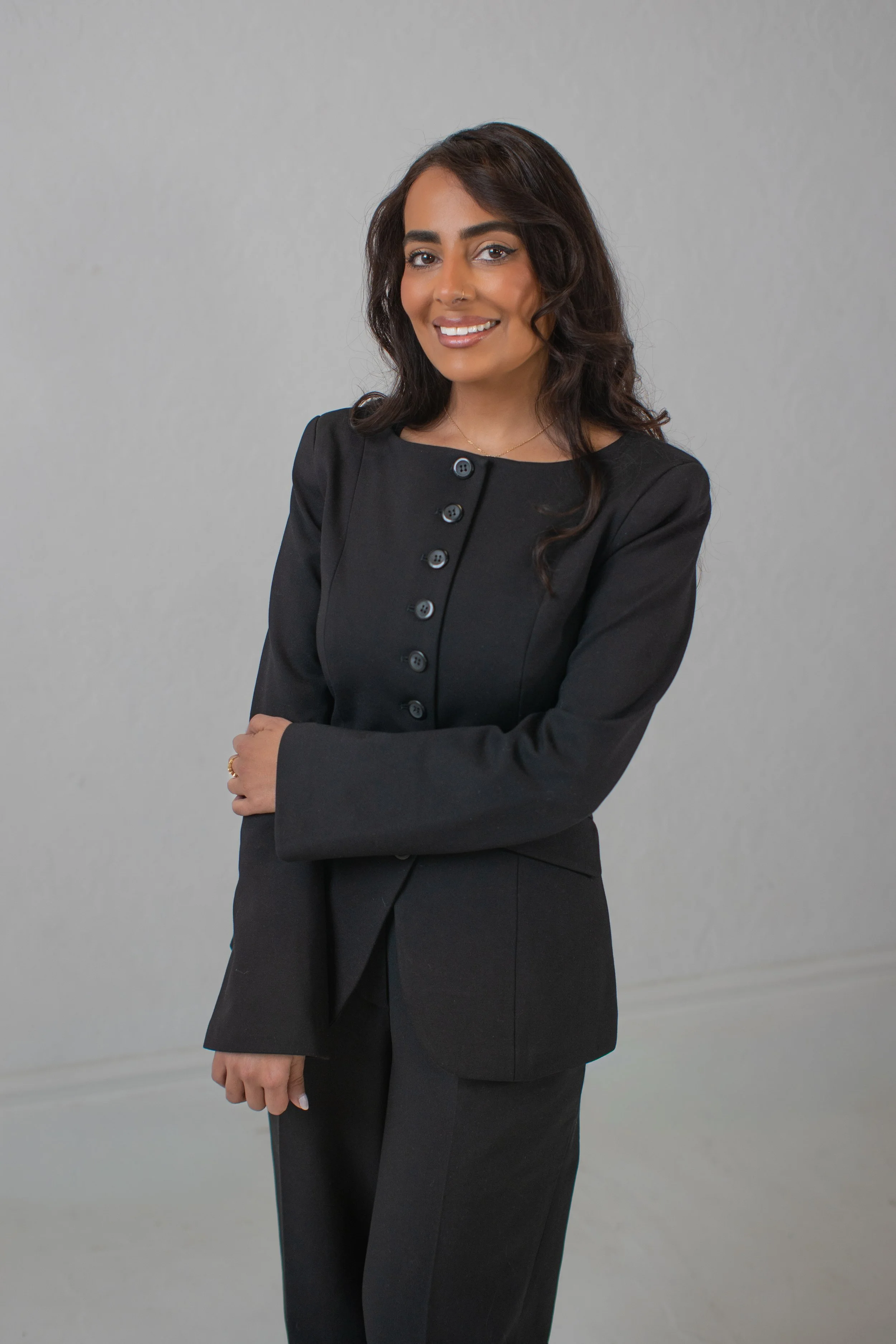A beautiful brown skinned woman stands confidently smiling in front of a grey backdrop wearing a black women's suit.