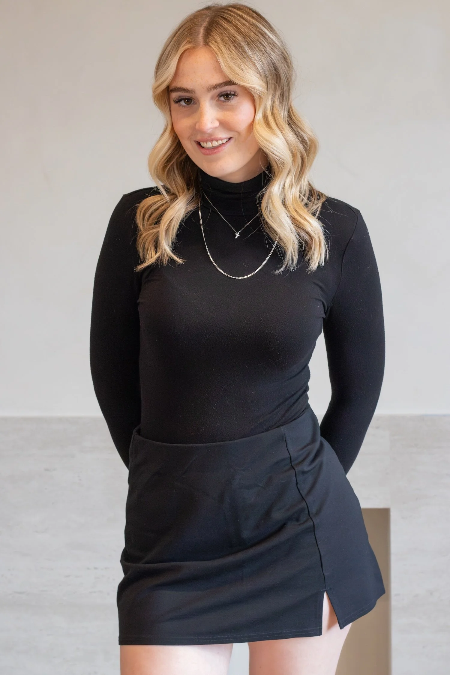 A beautiful, young blonde haired women stands smiling in front of a beige backdrop as she wears a black long sleeve turtleneck.