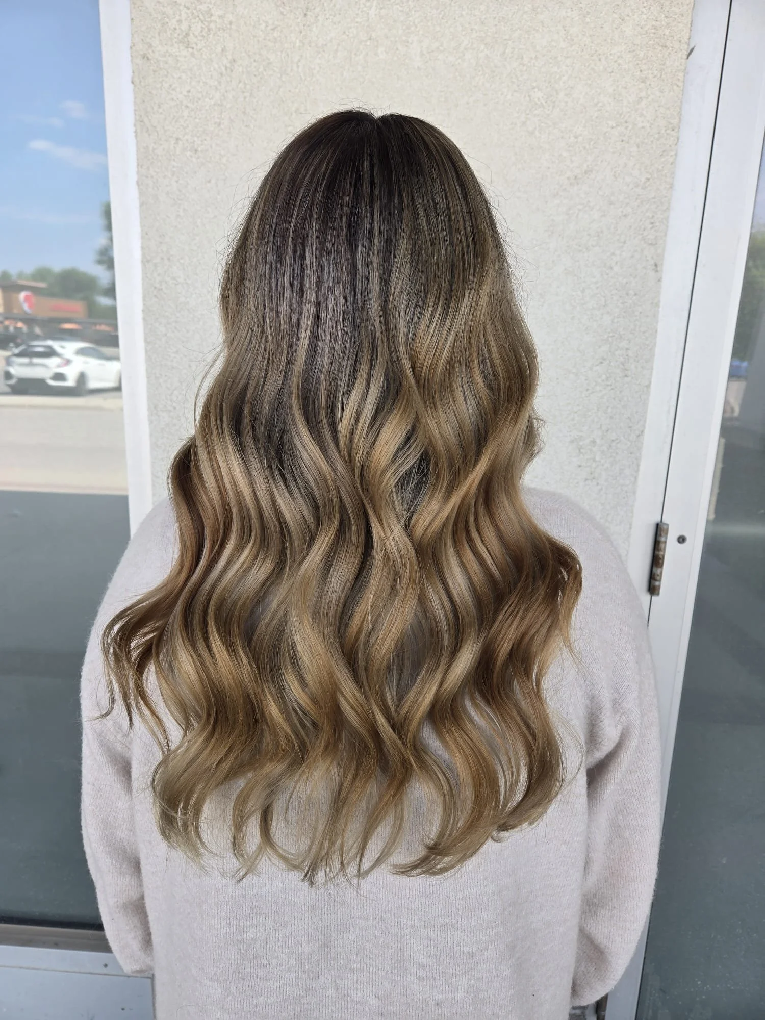A girl stands with her back to the camera, as her long, curly, and freshly balayge styled hair falls down her back.
