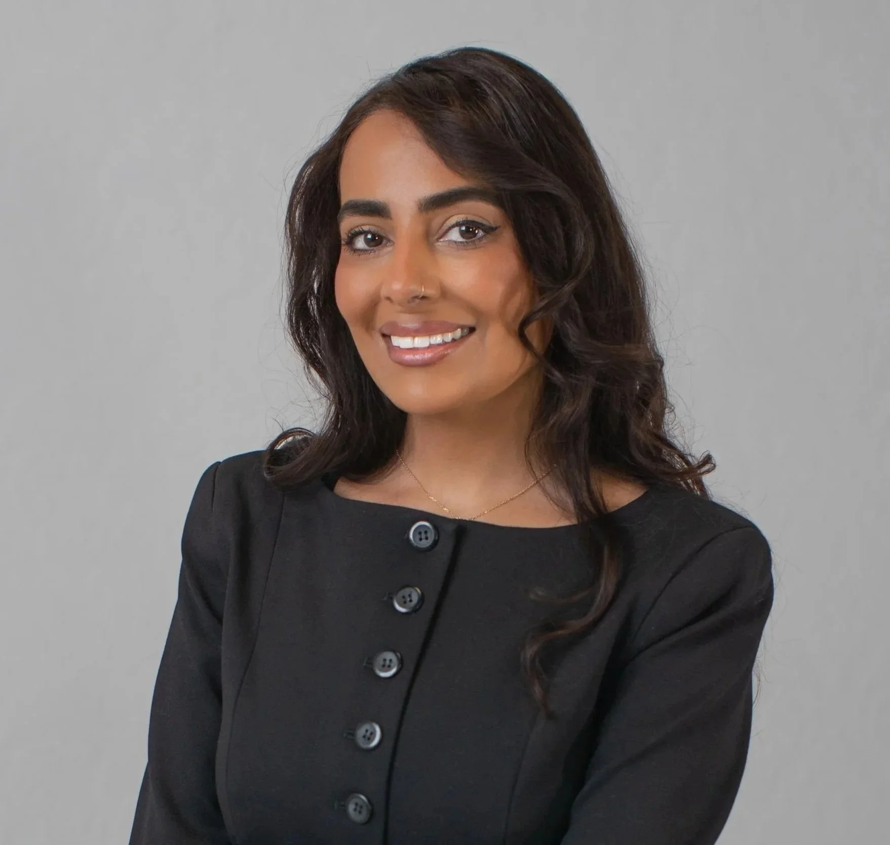 A confident, young, and brown skinned woman wearing a black button up suitstands smiling in front of a grey backdrop