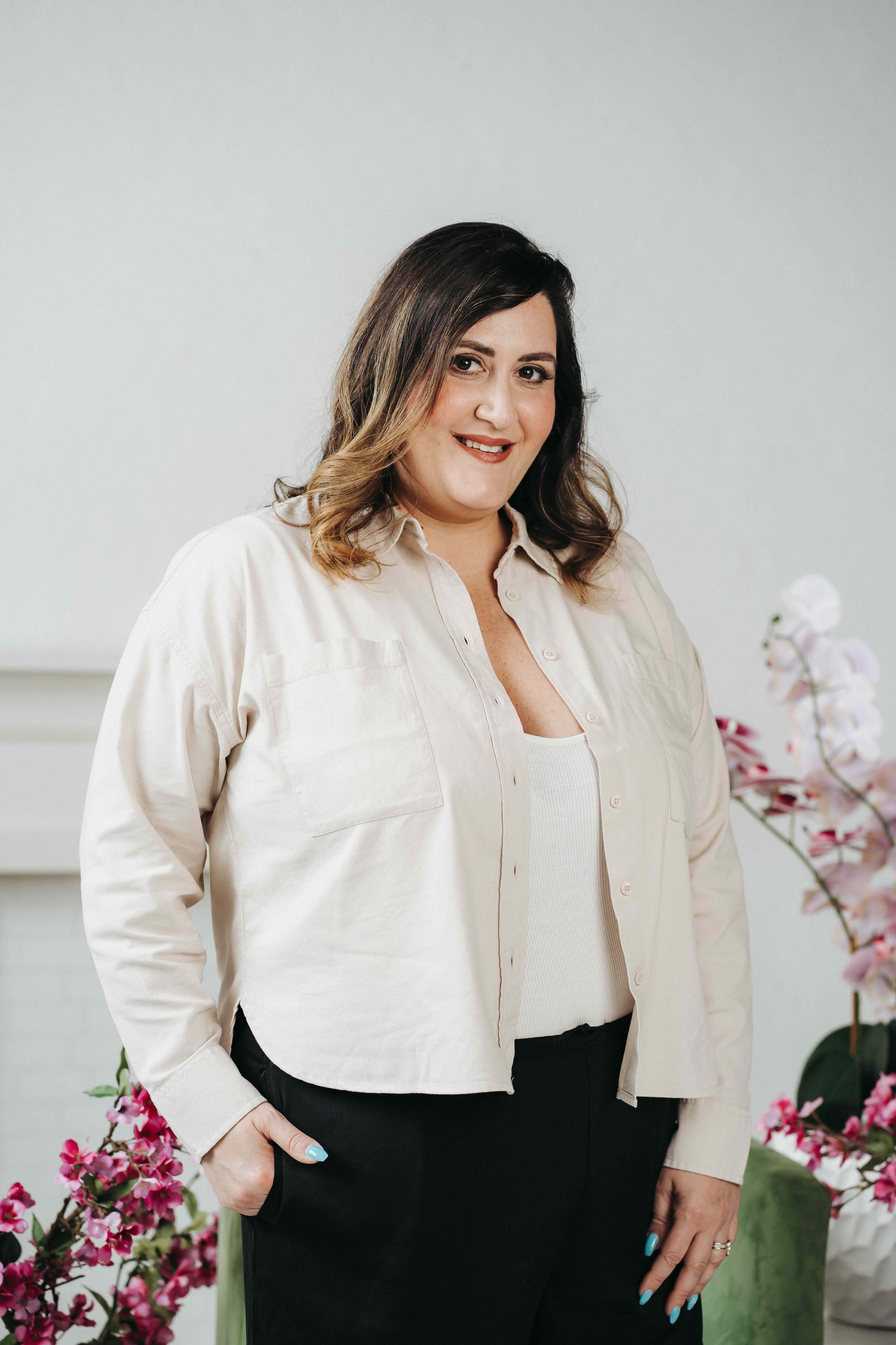 A white woman in her 40s stands confidently smiling in front of a grey backdrop.