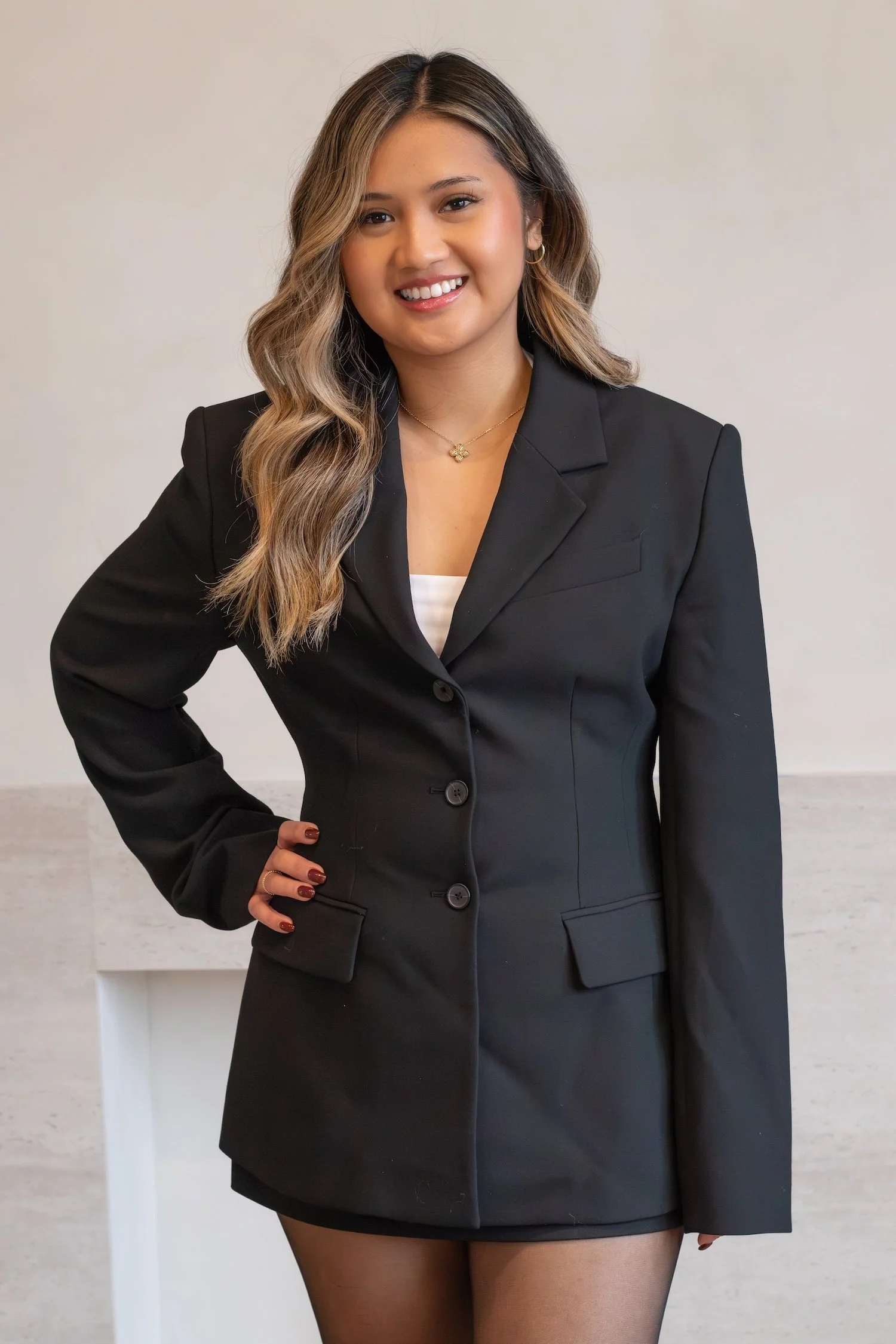 A beautiful filipino women with dark hair stands confidently in front of a beige background wearing a black blazer.