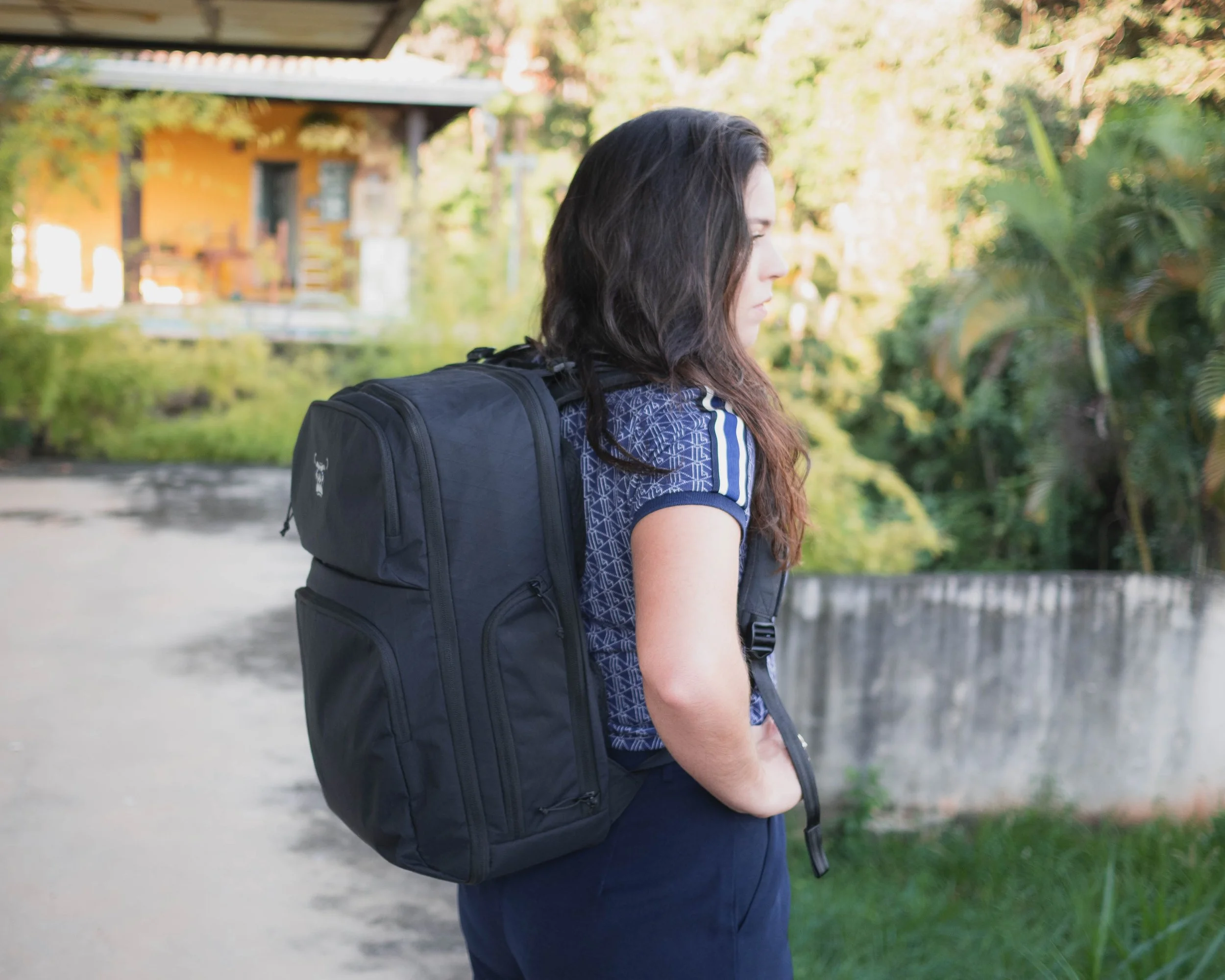 A woman with long dark hair wearing a blue patterned shirt and navy pants, carrying a black backpack, standing outdoors near trees and a yellow house with a gray roof, looking to the right.